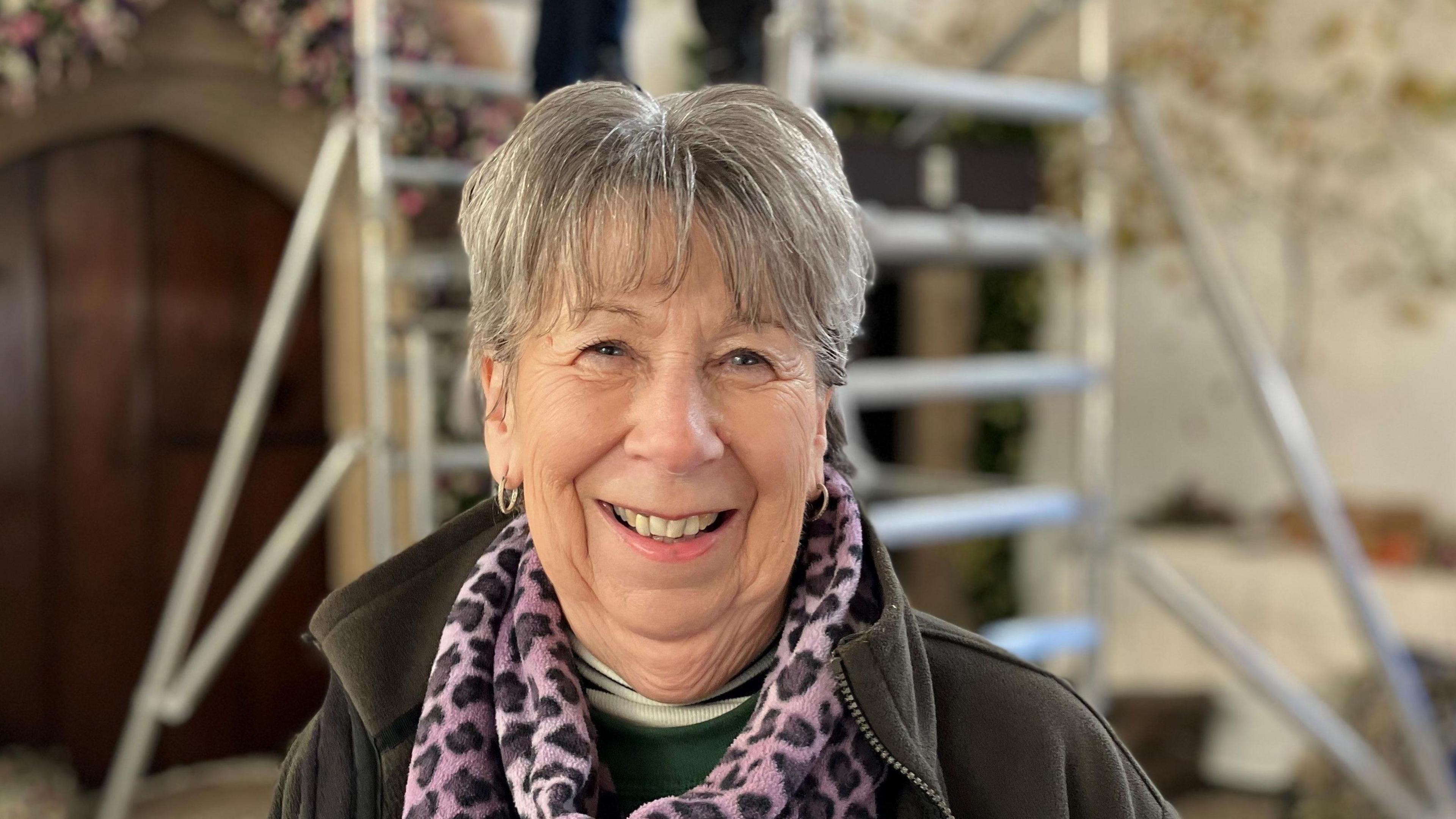 Lady with grey hair smiling at the camera. She is wearing a pink leopard print scarf and a grey fleece. 
