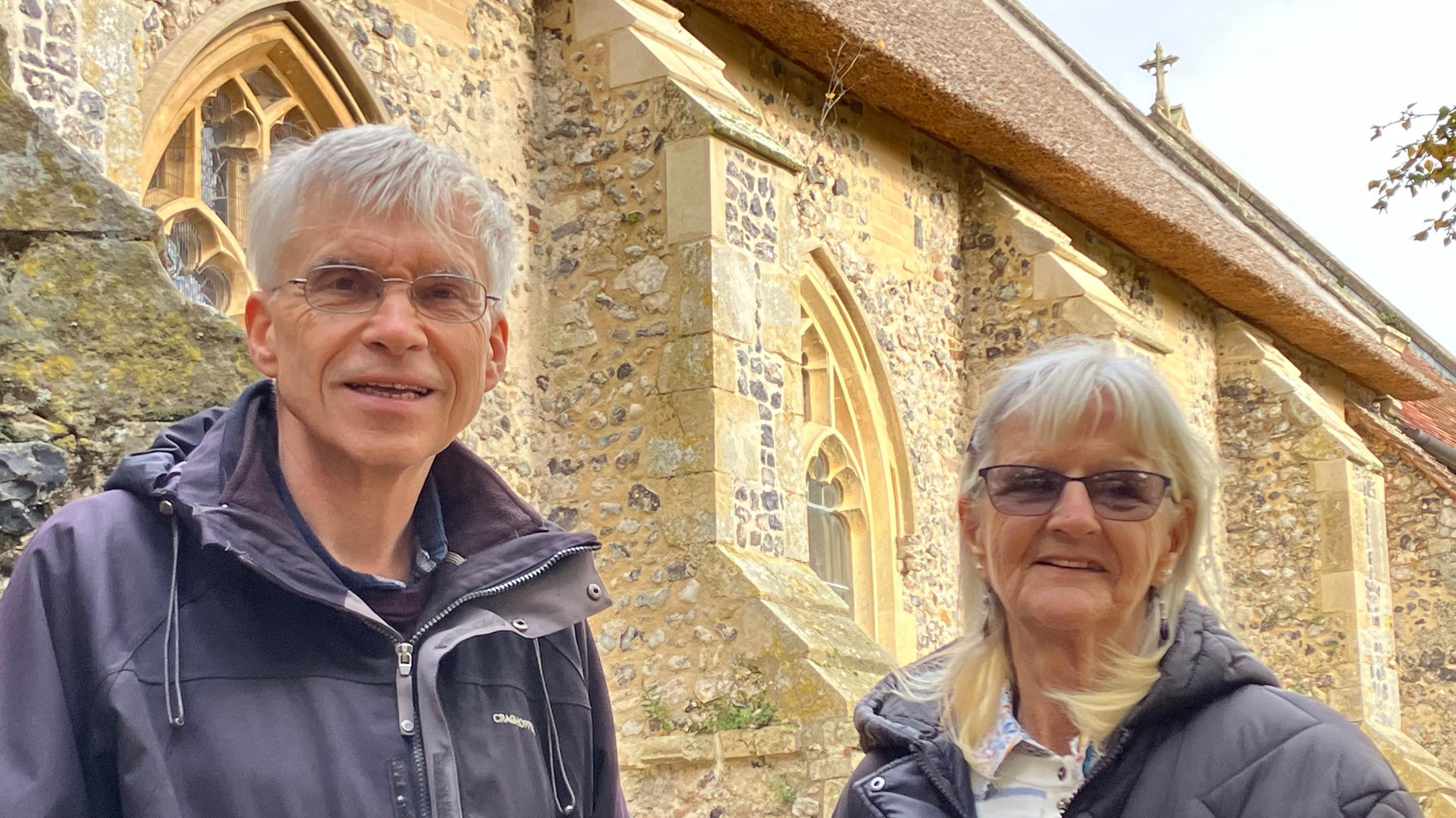 Ian Myhill, wearing glasses and a blue jacket, and Helena Lord, wearing glasses and a dark coat, stand together in front of the stone buttresses of the church