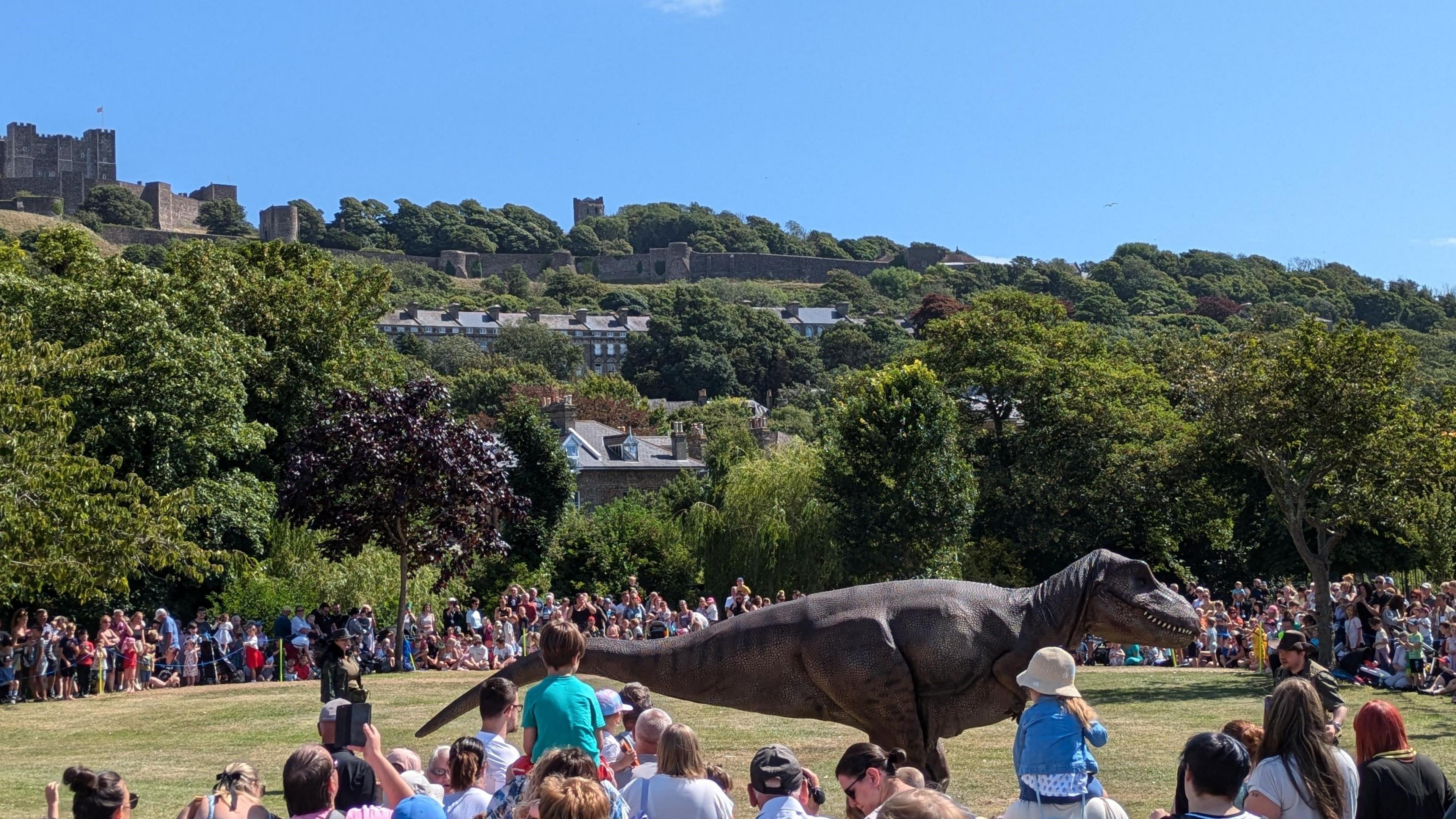 A dinosaur model in a field surrounded by adults and children.