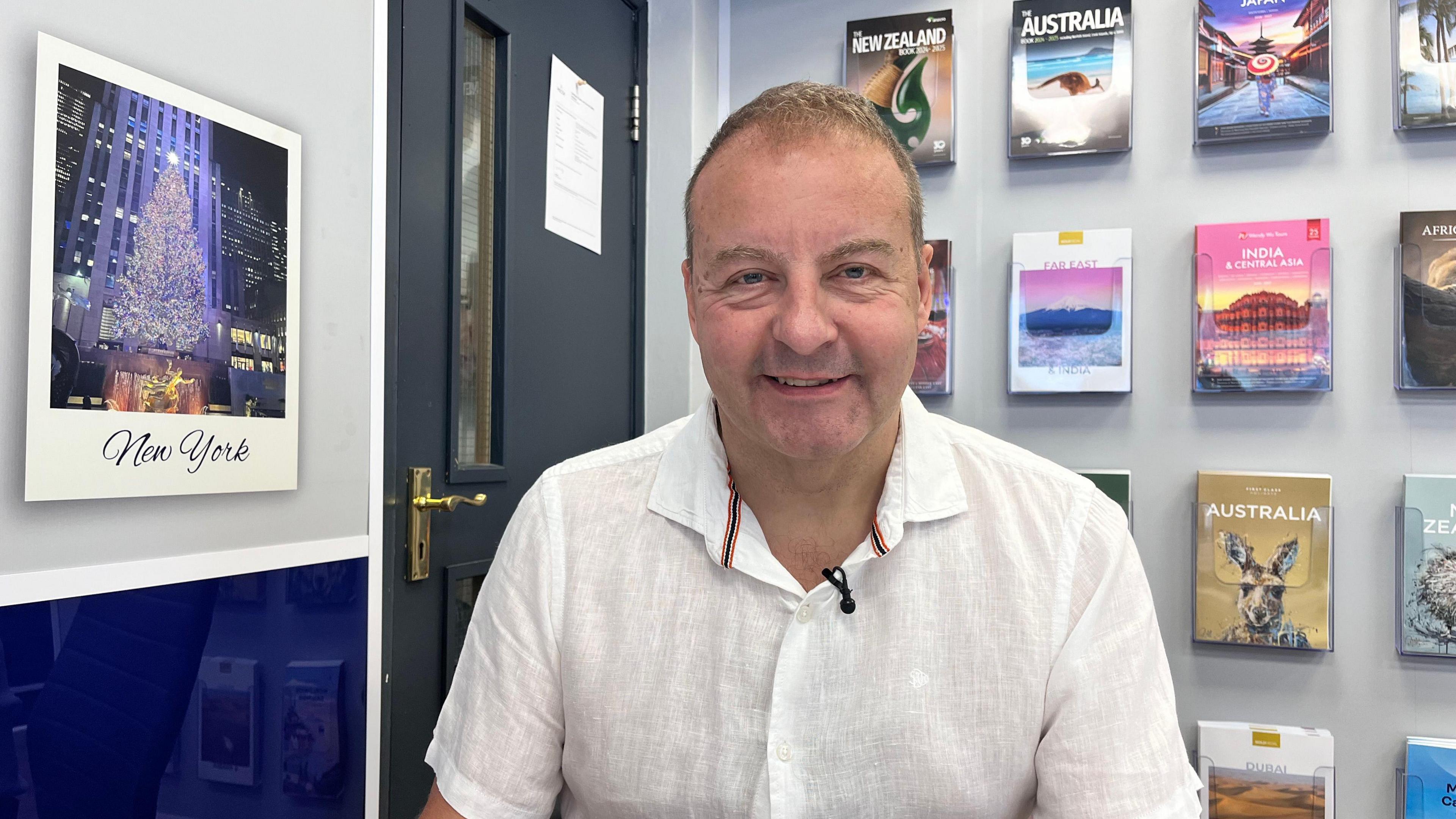 Andrew Earle sits in front of his computer in his travel agents in Hull city centre wearing a white shirt with holiday brochures to various destinations in the background.