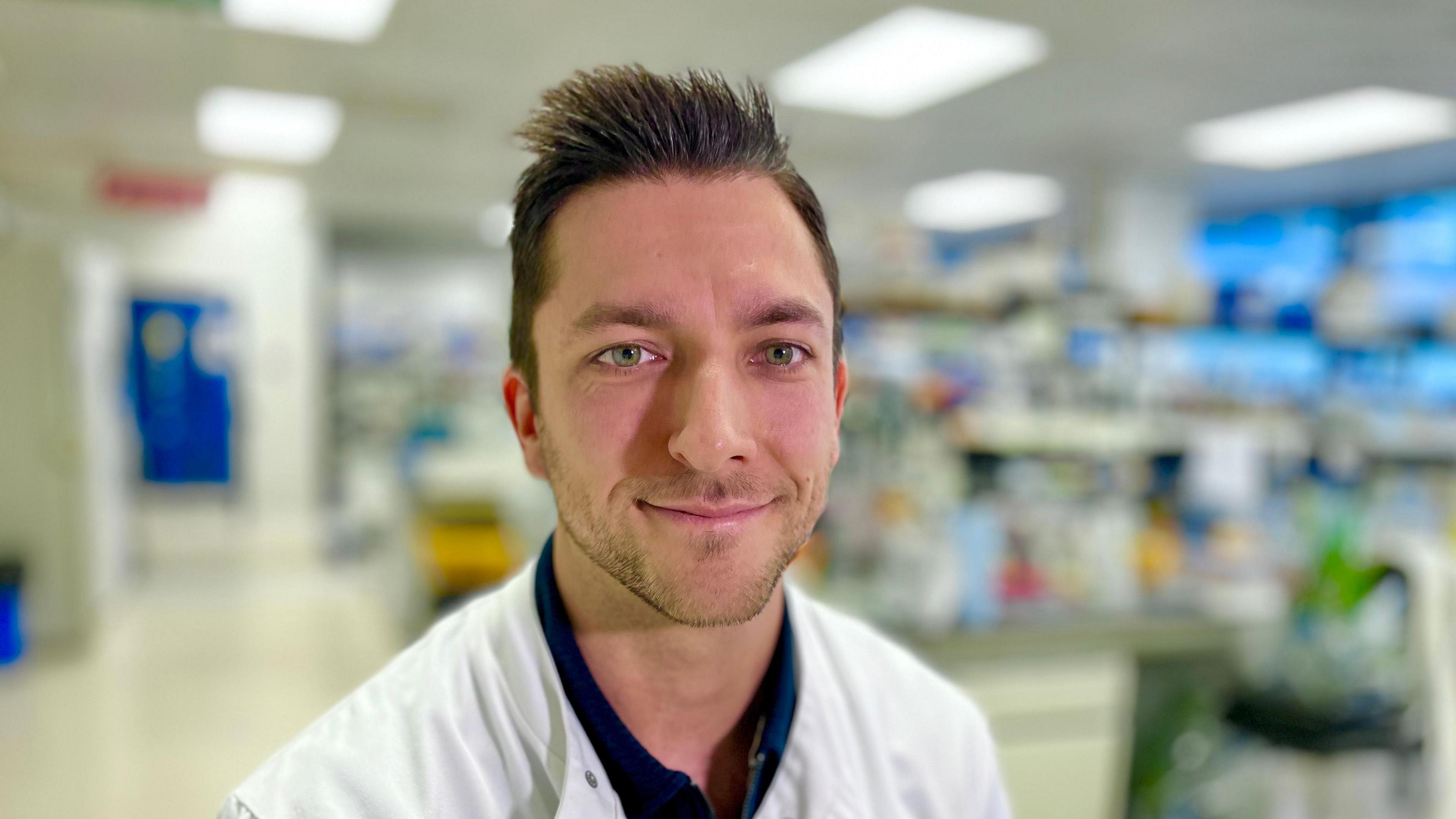 Damien Caubriere smiles at the camera. He has short, spiky brown hair and is wearing a white lab coat over a dark-coloured shirt. He is standing in a laboratory.