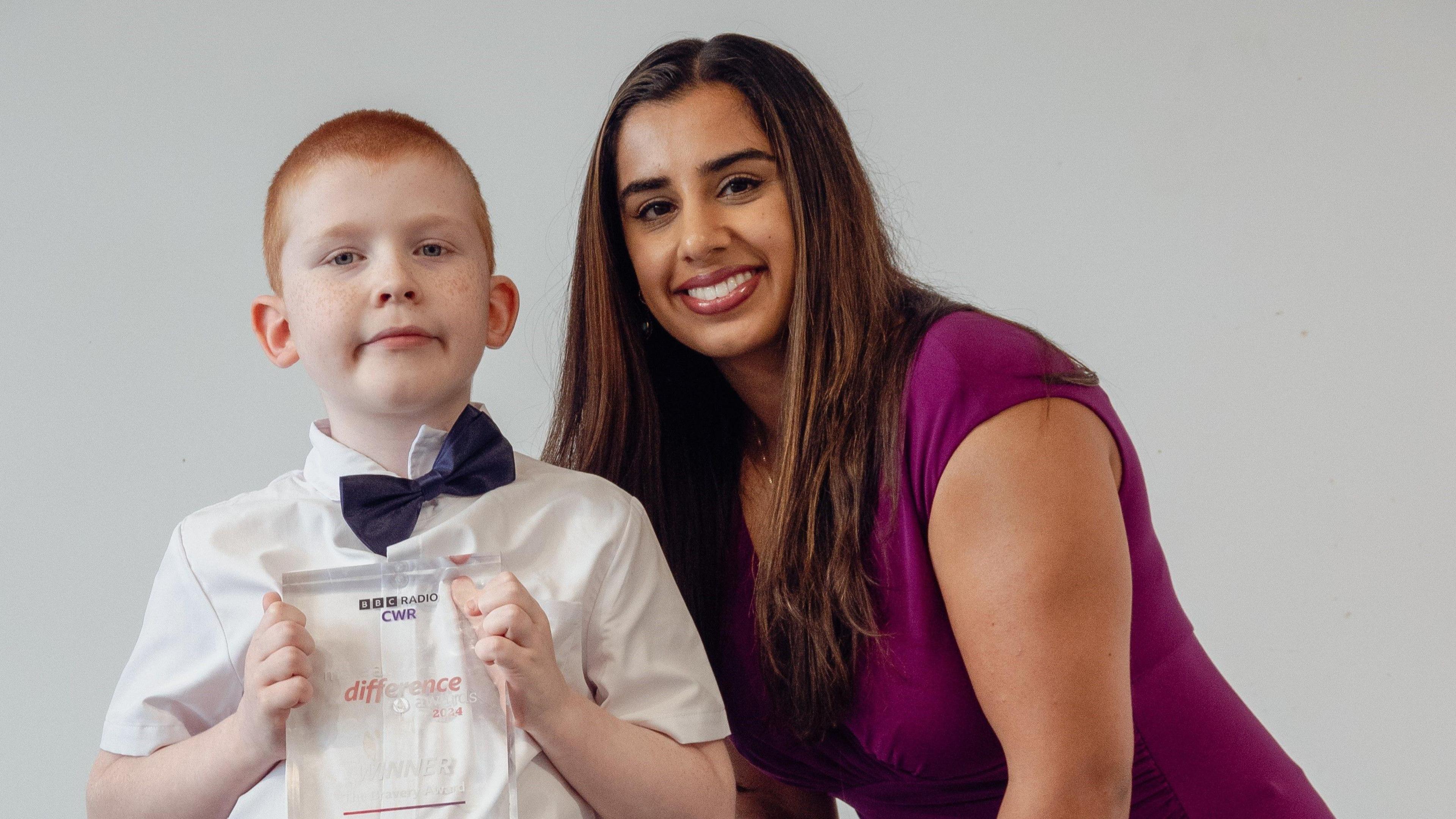 A young boy with ginger hair is holding a glass award. He is wearing a white shirt and blue bow tie. A woman with brown hair is standing next to him, wearing a plum coloured dress.