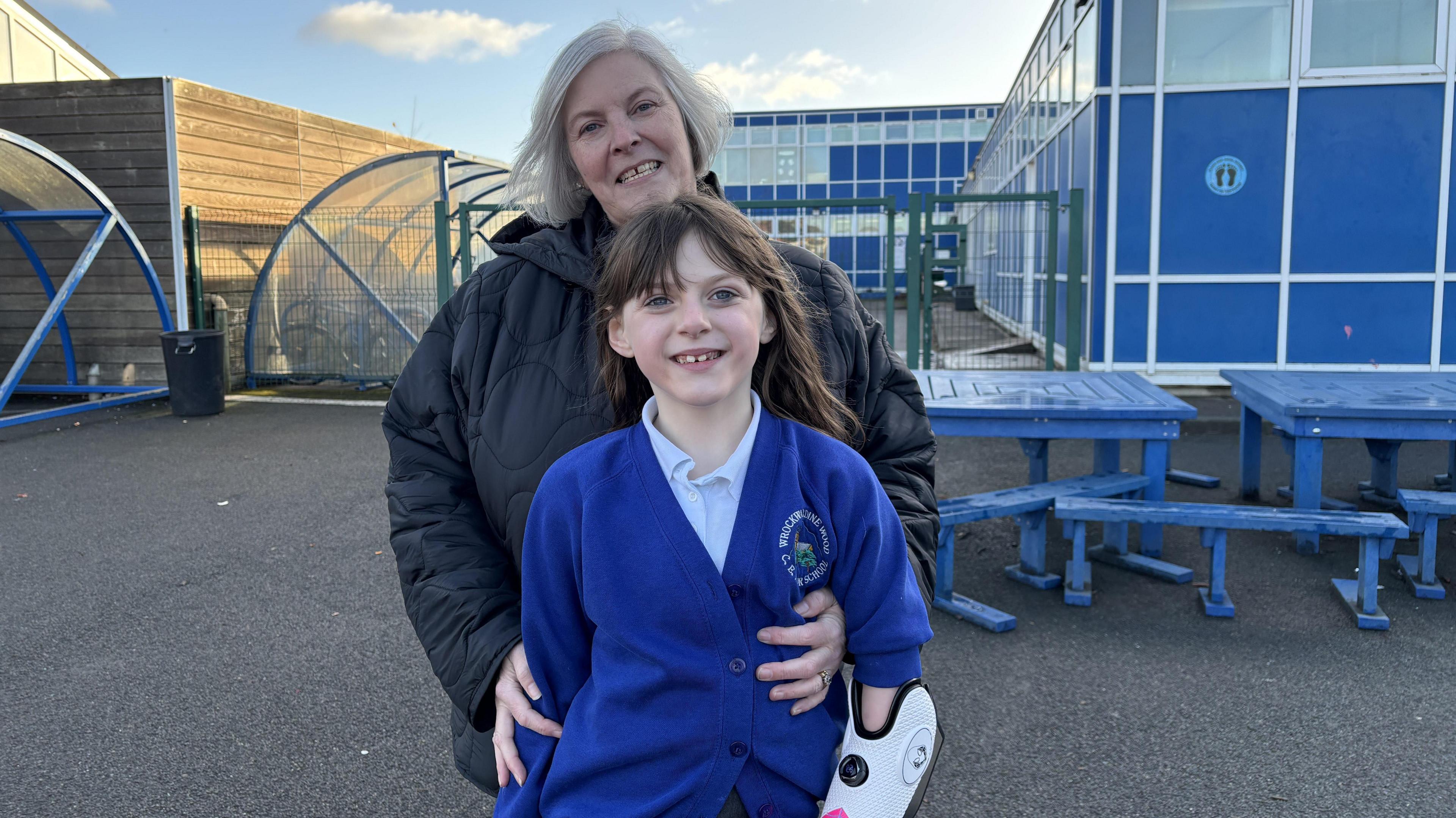 Zoey is in her blue school uniform and is smiling at the camera. She has brown hair and a fringe. Part of he bionic arm can be seen as the sleeve of her cardigan has been pushed up. The bionic arm has a white casing and black outlining. Her grandmother is hugging Zoey from and has her arms wrapped around her waist. The grandmother is wearing a black puffer coat and is smiling into the camera. Behind them is Zoey's school, which is a blue block with rectangular windows. To the left of them are bike sheds with bike racks in them and there are blue outdoor metal tables and benches on the right. 