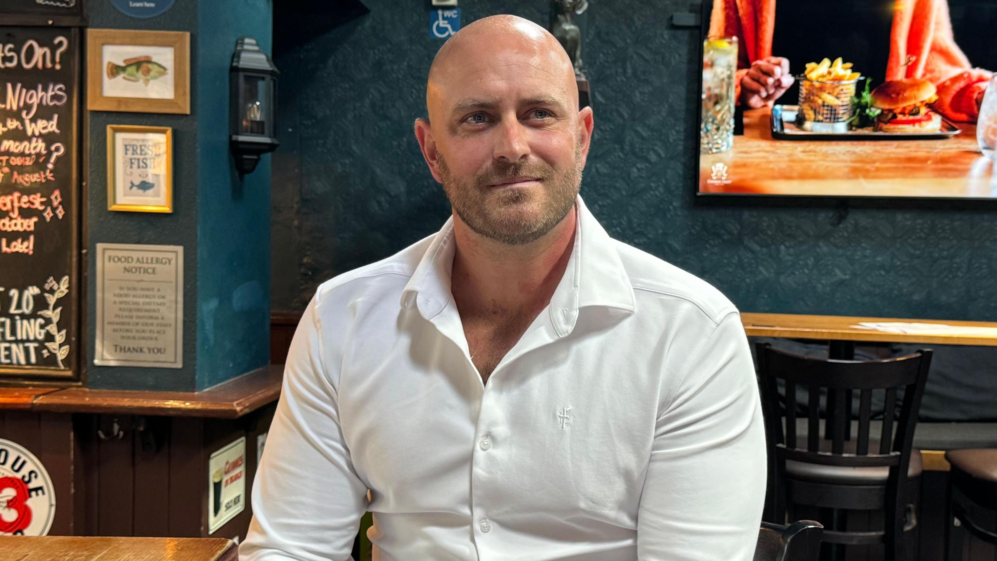 Dave Burns is sitting on a stool in the bar of the pub. He is wearing a white collared shirt. A television screen is in the background