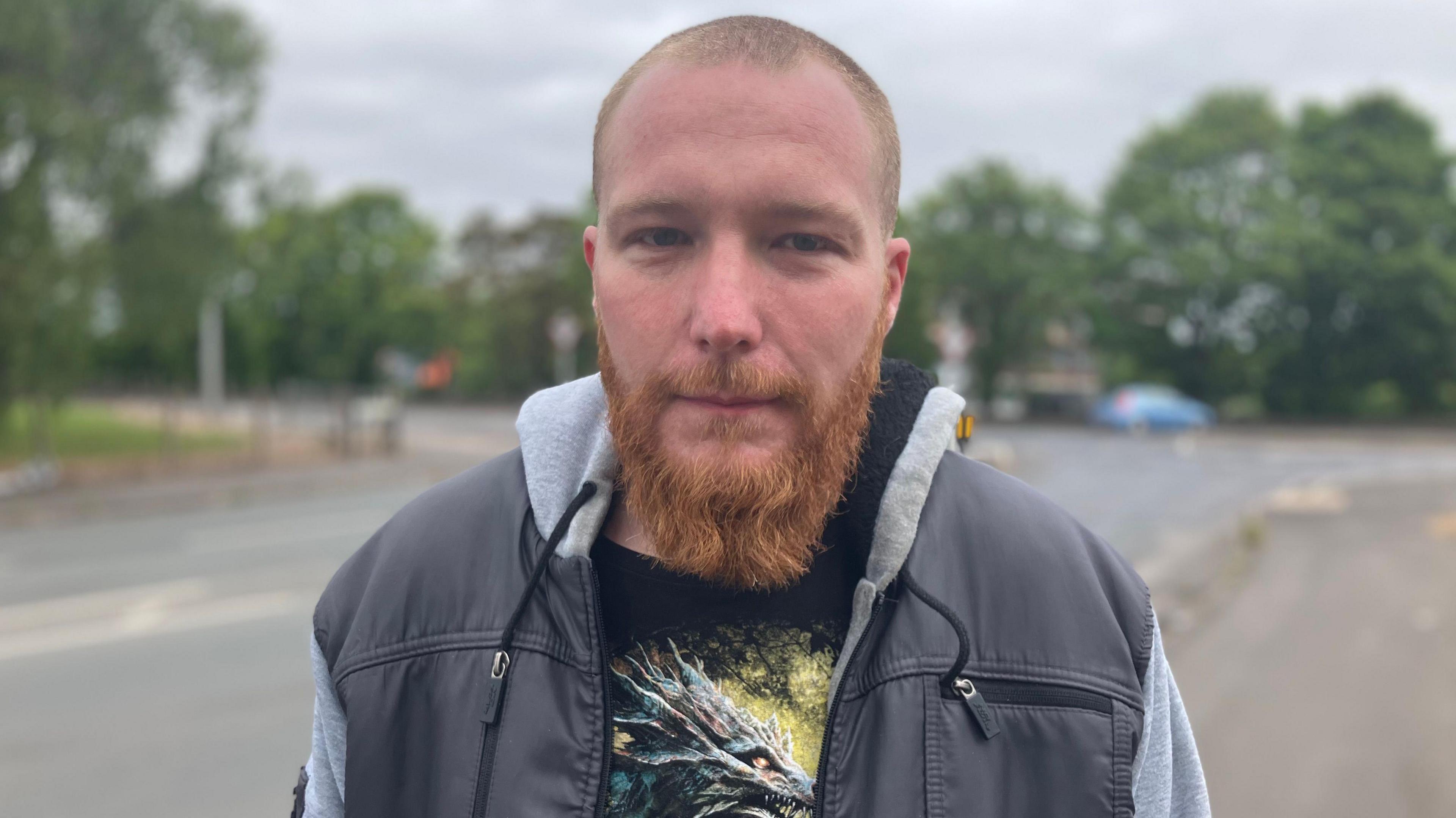 John stands on the side of the road, wearing a t shirt and hooded jacket, looking straight into the camera.