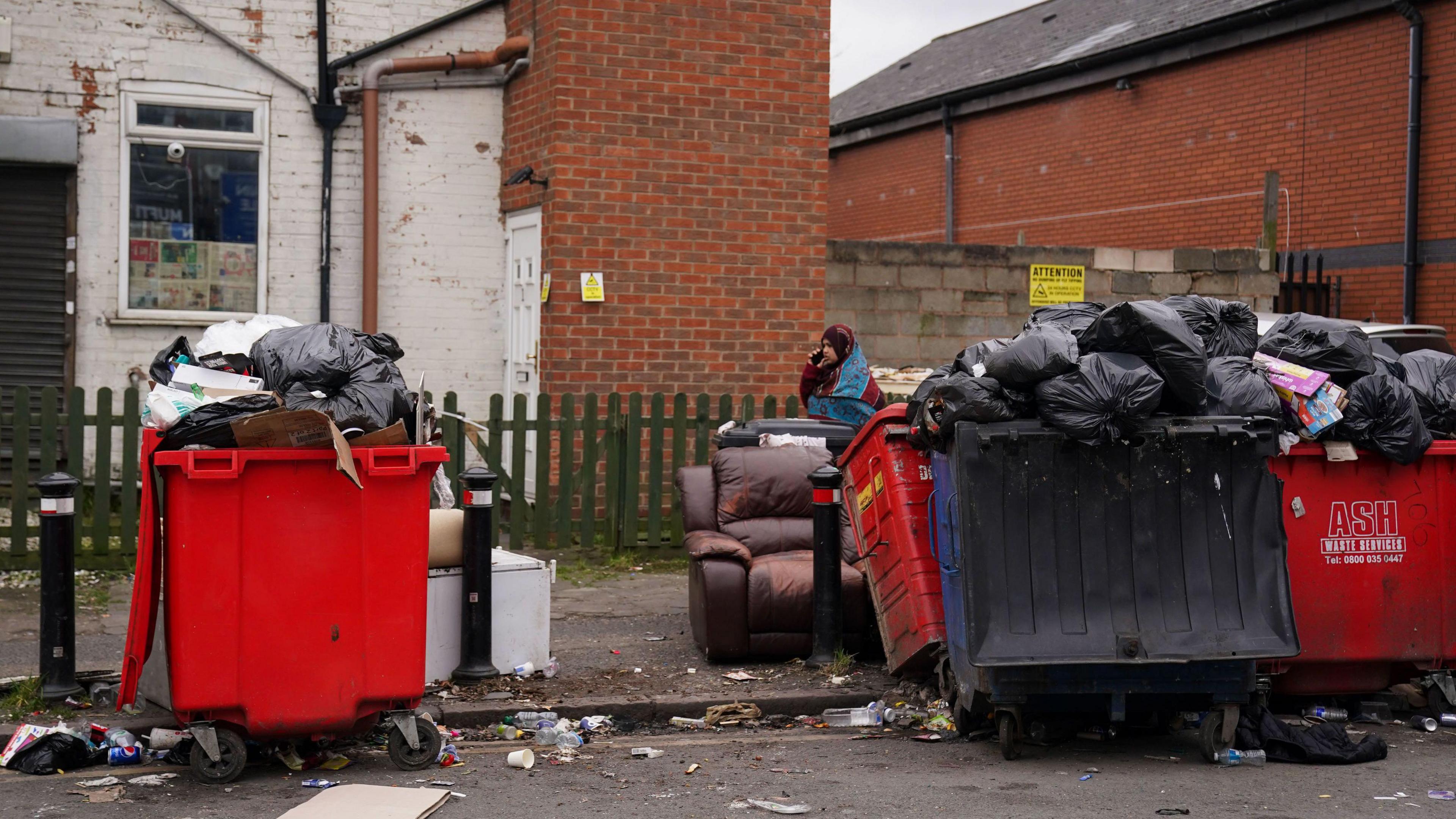 Uncollected refuse bags in Birmingham in March. Red bins are overflowing. There has also been some fly-tipping. It looks a mess. A woman is walking past talking on her phone.