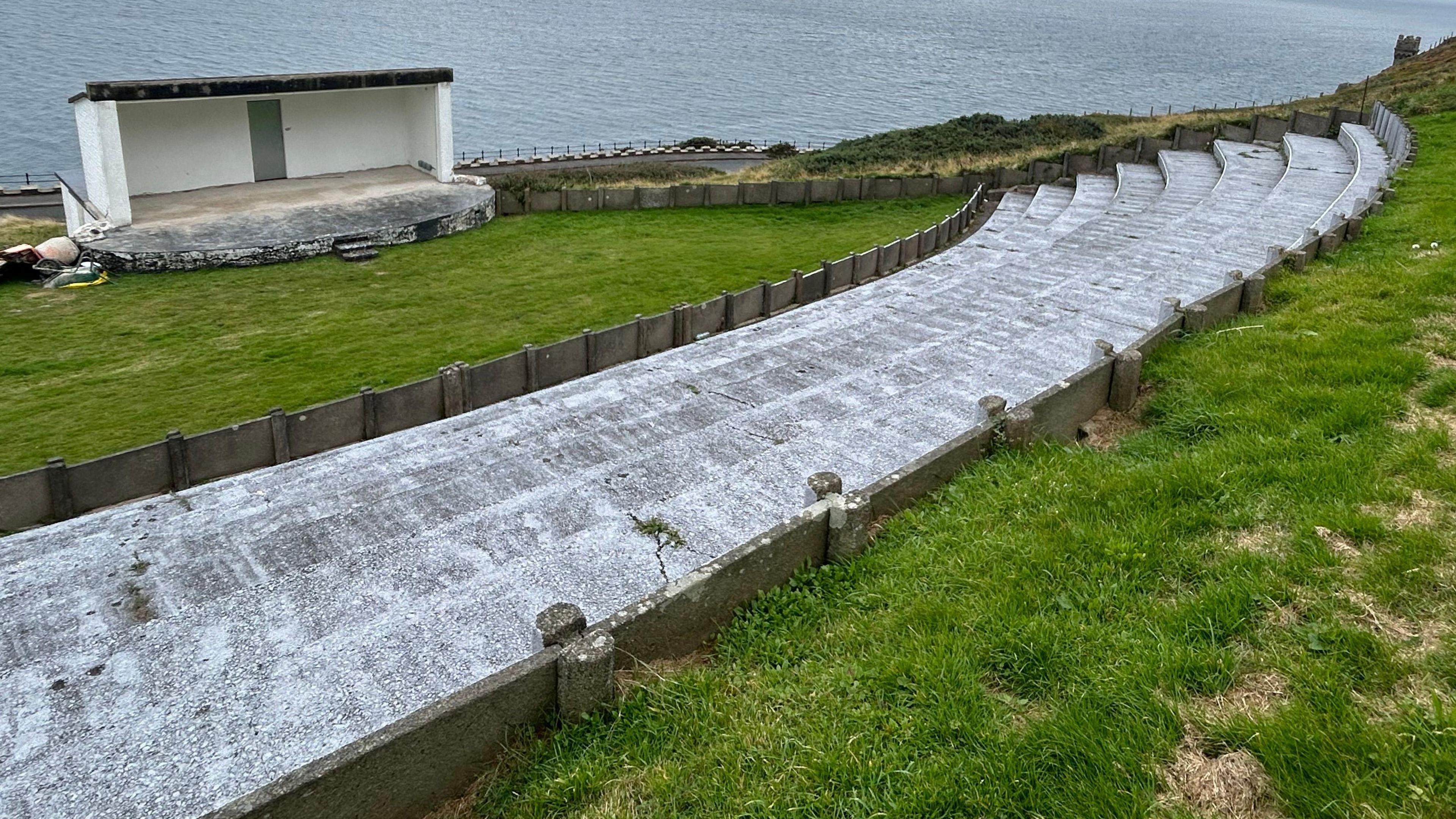 The Douglas Head amphitheatre, which has a sheltered performance space with three walls and a raised stage and the sea in the background. There is a stretch of grass in front of it, with a semicircle of concrete steps sitting in a grassy bank.