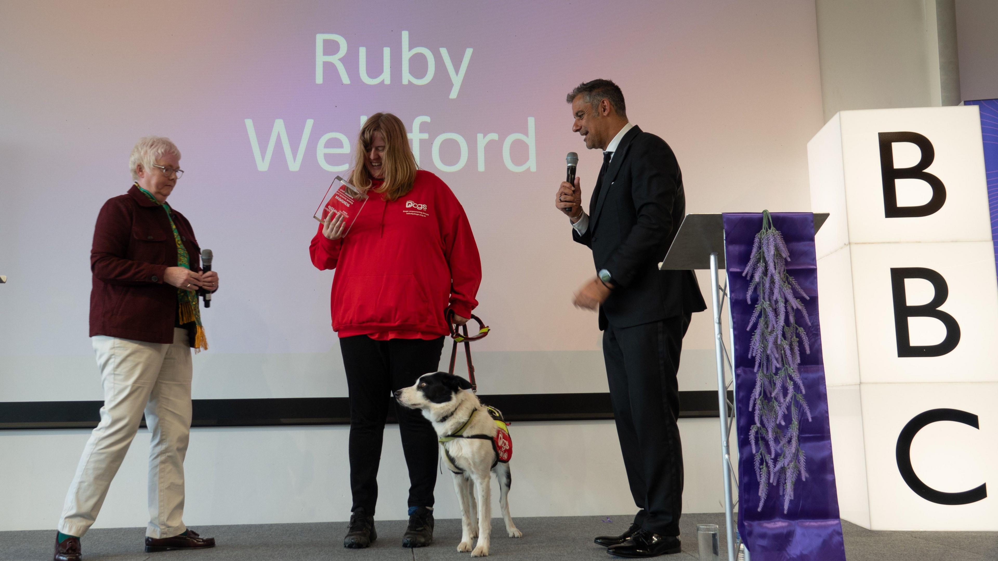 A woman stands on the left of a stage with a microphone. In the centre, is a woman in a red hoodie holding a glass award. She has an assistance dog on a lead next to her. To her right is a man with a microphone. There is a purple plinth and a large light-up BBC sign