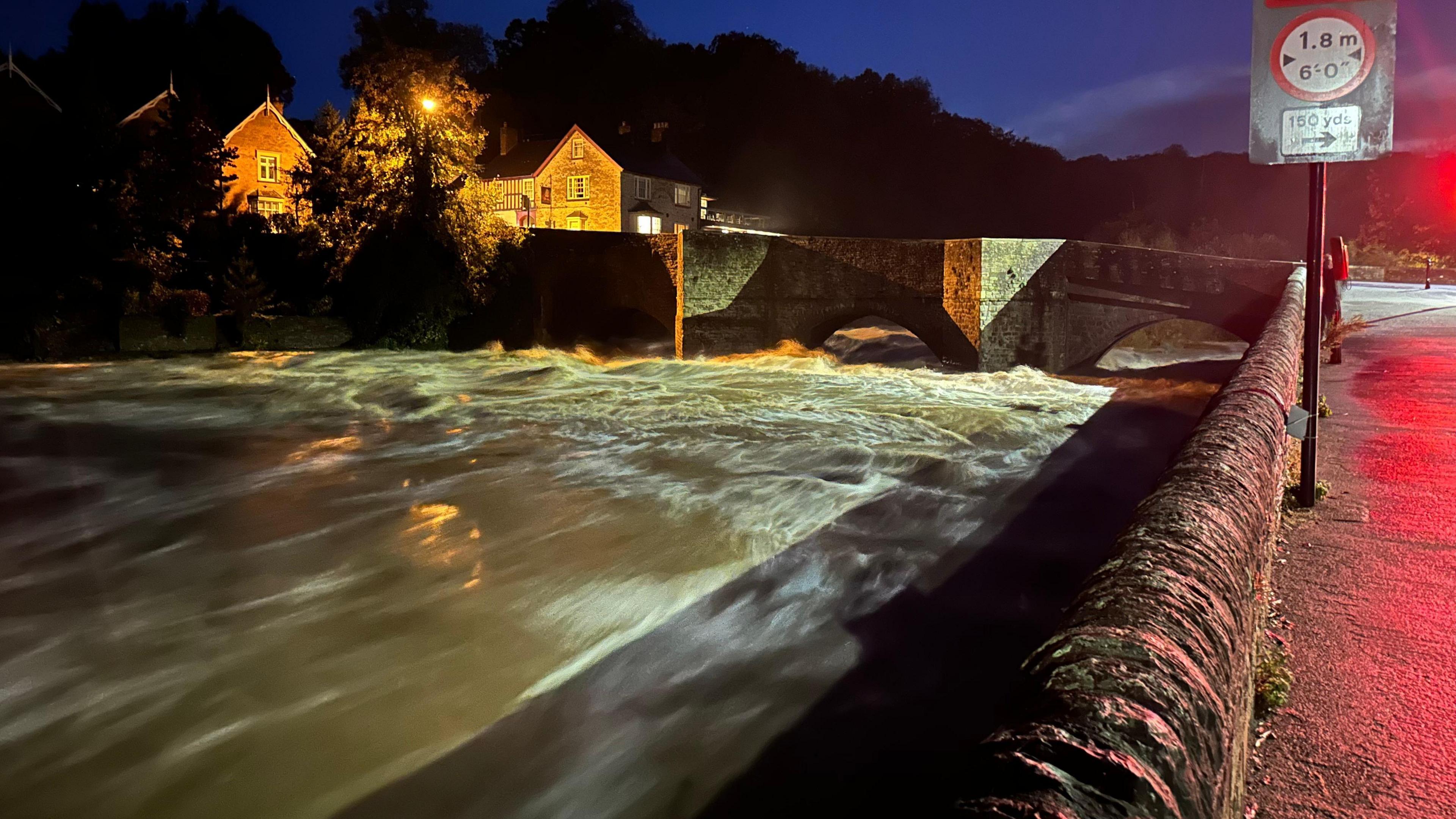 A fast-moving river, emphasised by a slight blur on the water's surface, rages at night under a stone bridge with a pub overlooking it in the background.