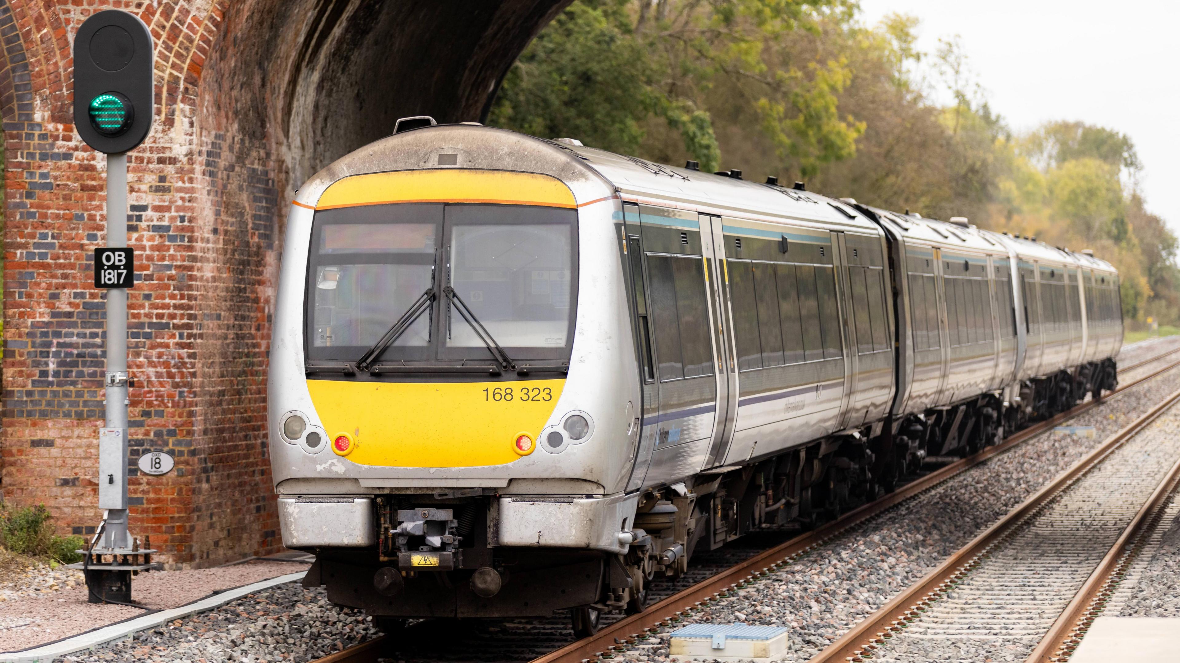 A test train going under a red brick bridge next to a railway signal which is lit green. The train is yellow and silver with two large windows at the front. The train is made up of three carriages.