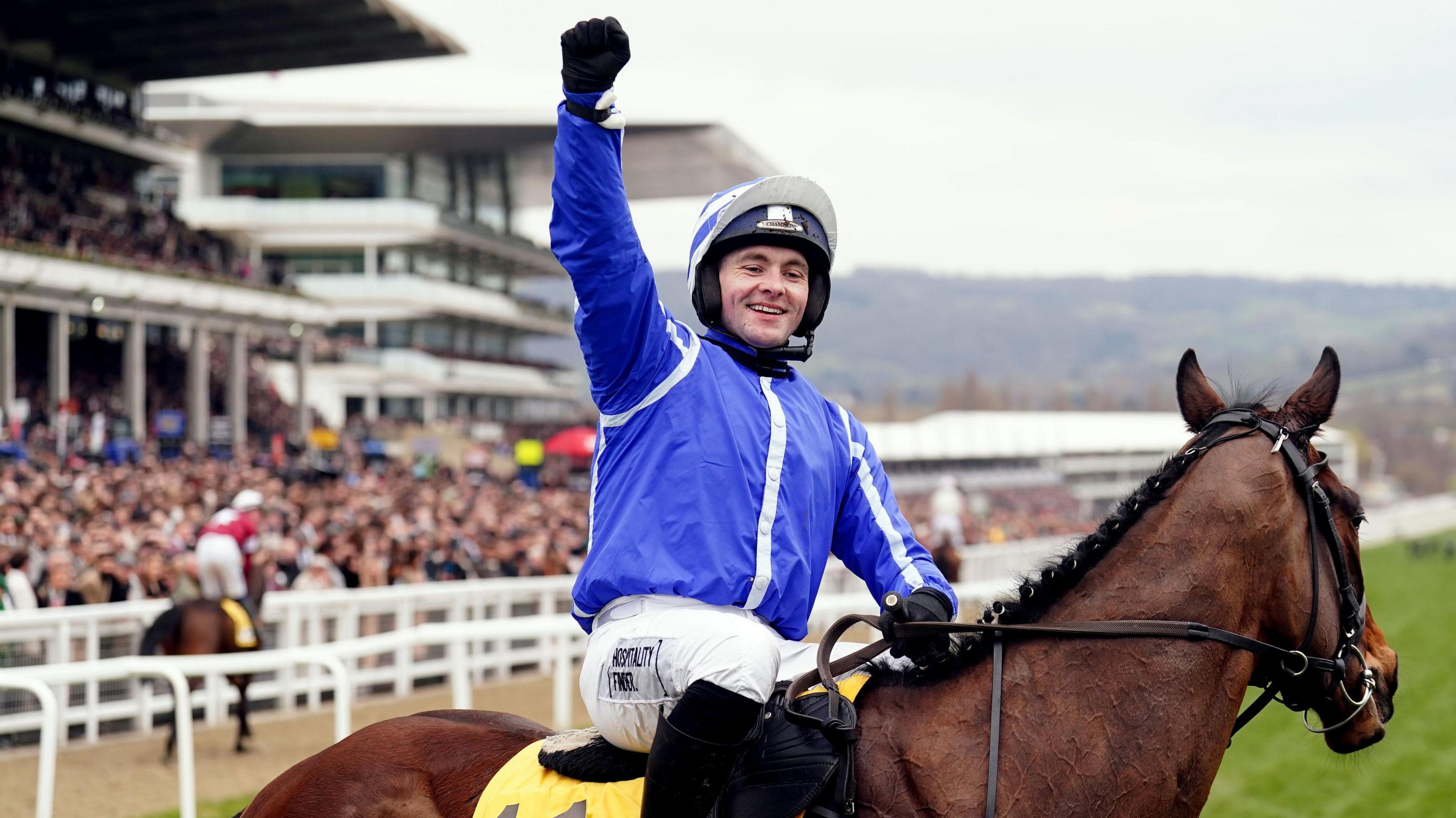 Jonjo O'Neil wearing a blue jacket, white slacks and a blue helmet, sits on horse Poniros with one arm raised in the air in celebration