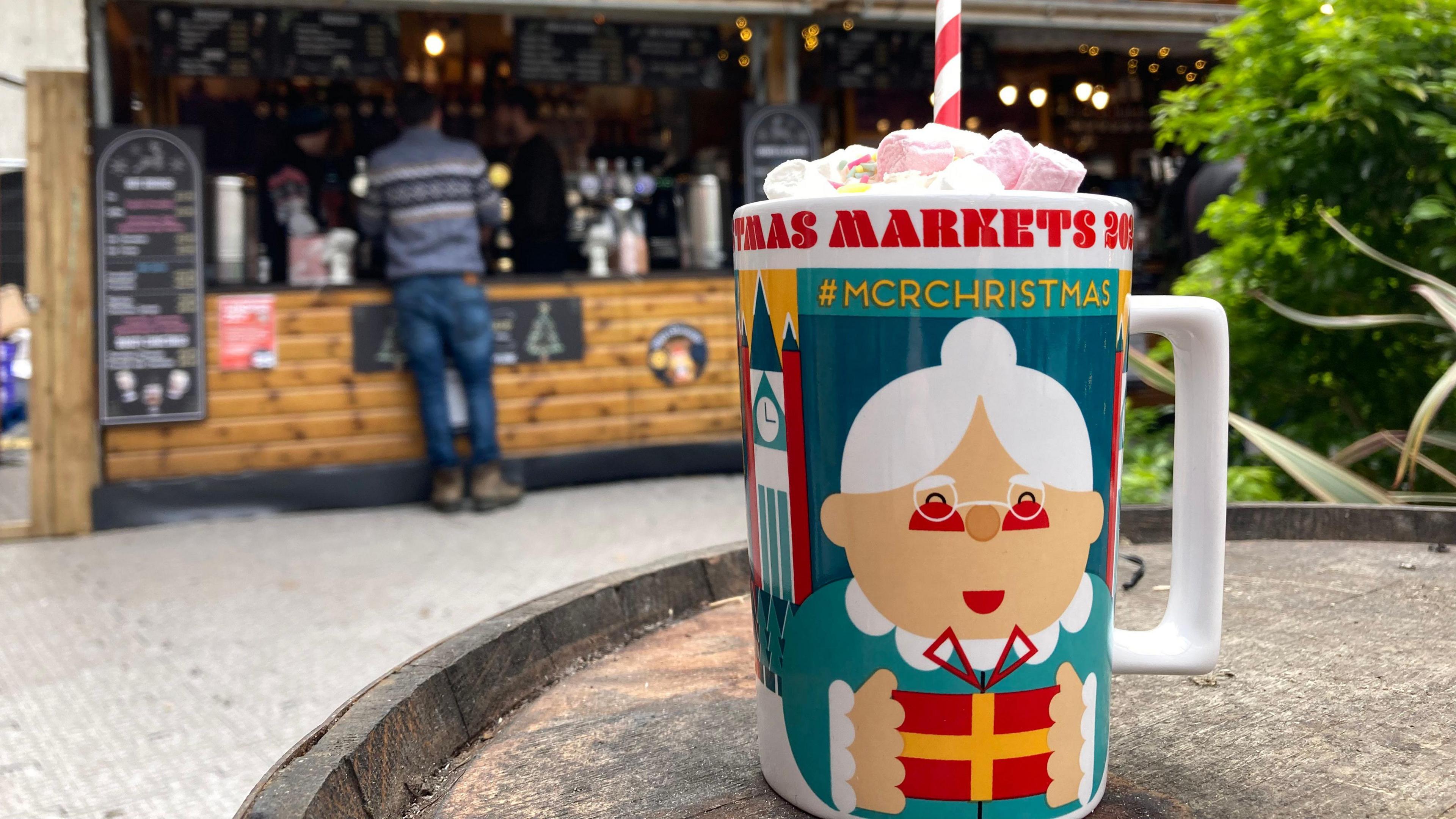 A mug bearing a decoration of an old lady sits on a table in front of a Christmas market stall 