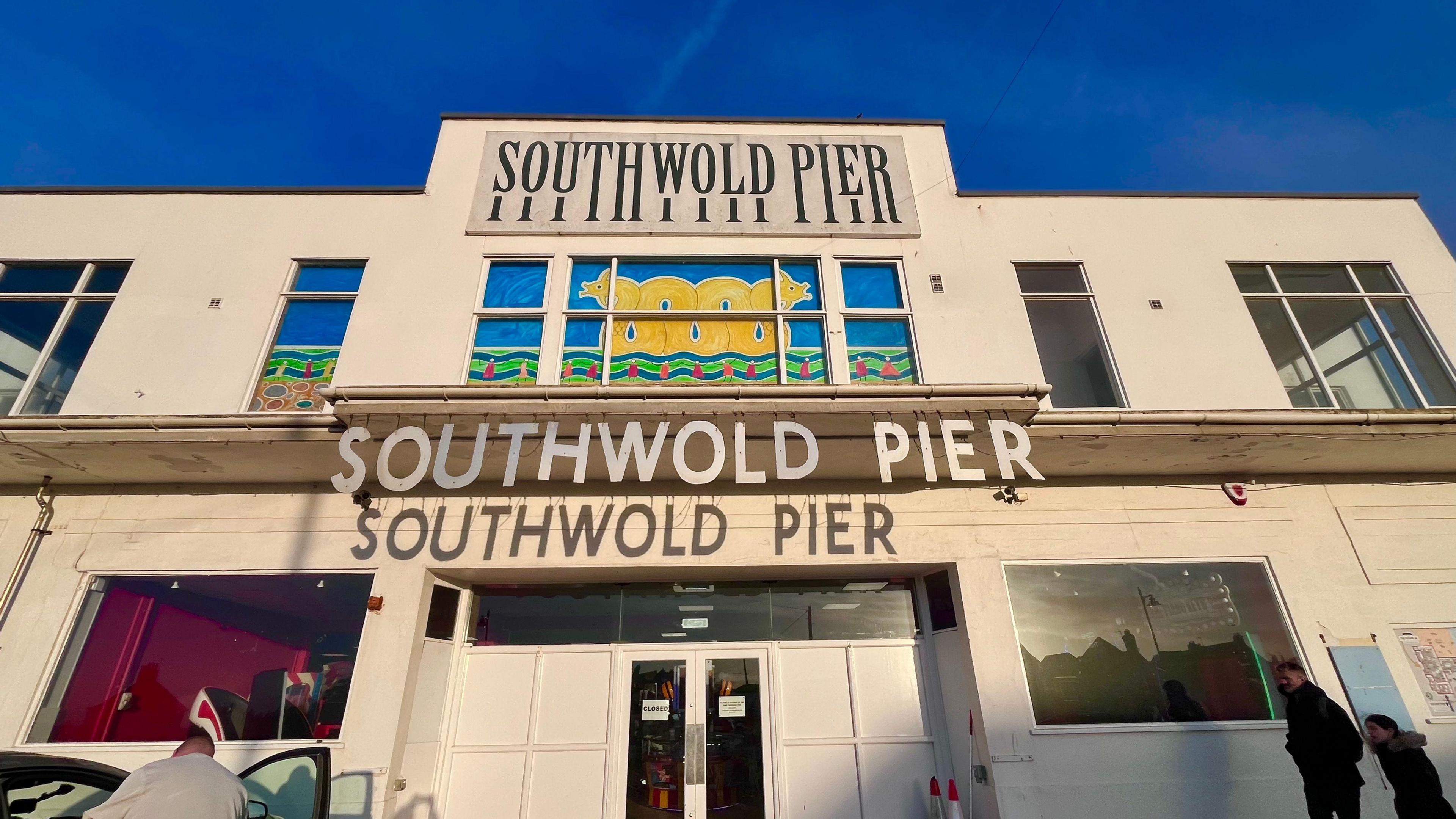 A building on the Southwold pier that is cream coloured with windows and doors, and two signs that read 'Southwold Pier' on it. People can be seen walking outside of the entrance. 