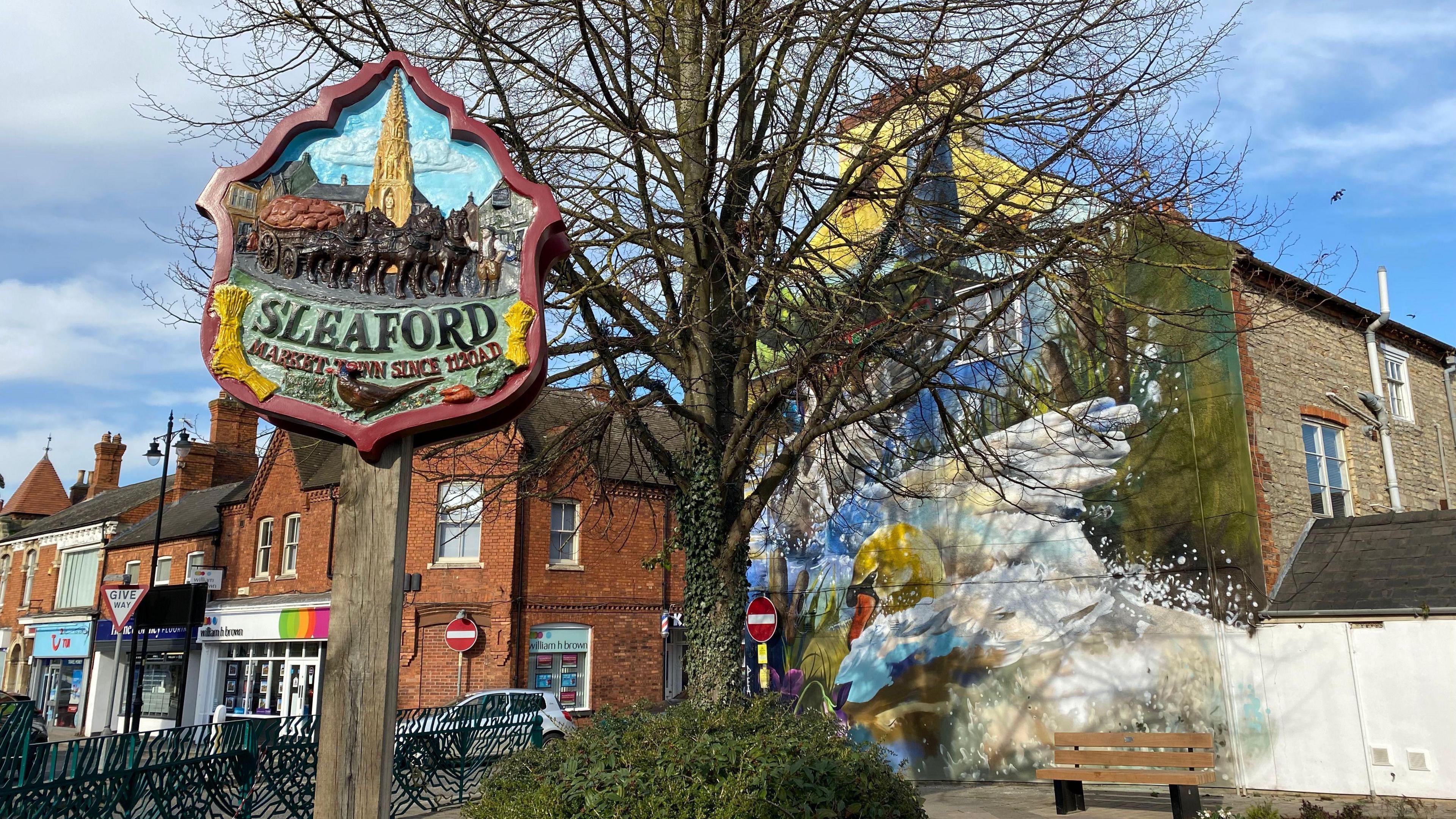 A wooden "Sleaford" sign in the foreground with the colourful mural behind a tree in the background.