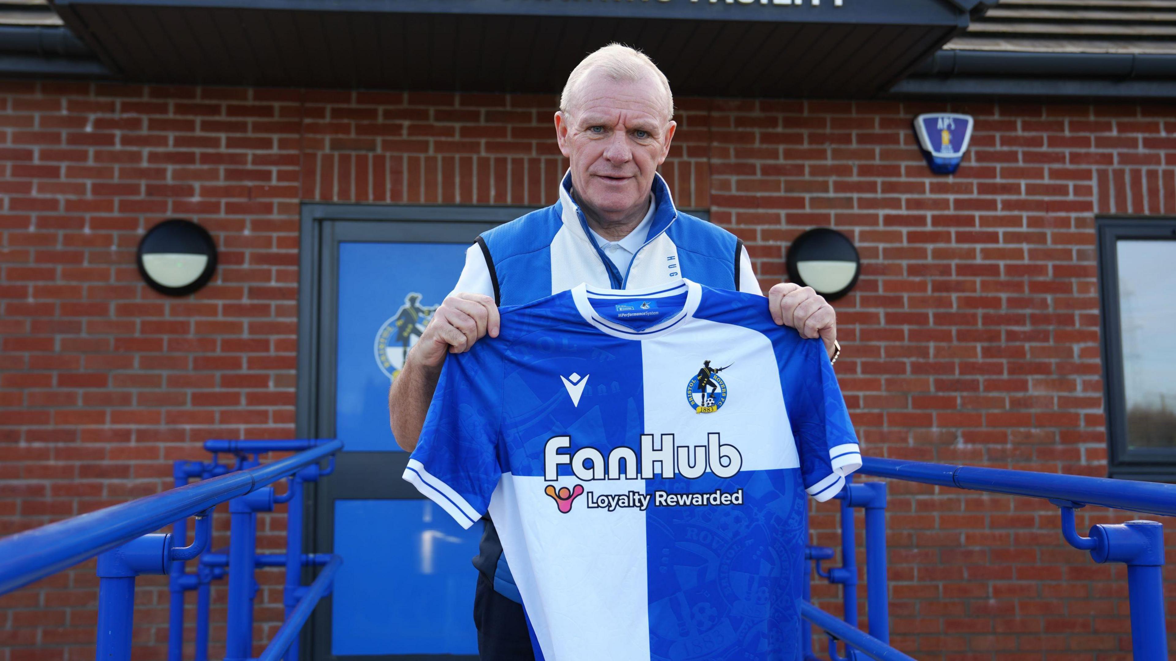 Steve Evans stands outside Bristol Rovers' training ground on a pathway holding a Bristol Rovers shirt in front of him