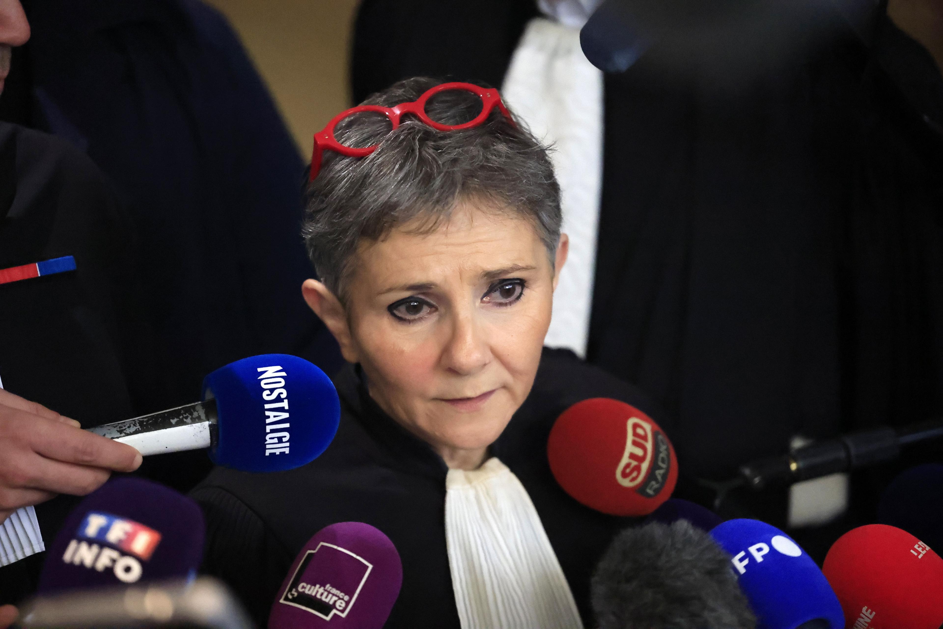 Béatrice Zavarro with grey hair and red-rimmed glasses on her head stands in front of several microphones as she speaks to the press.