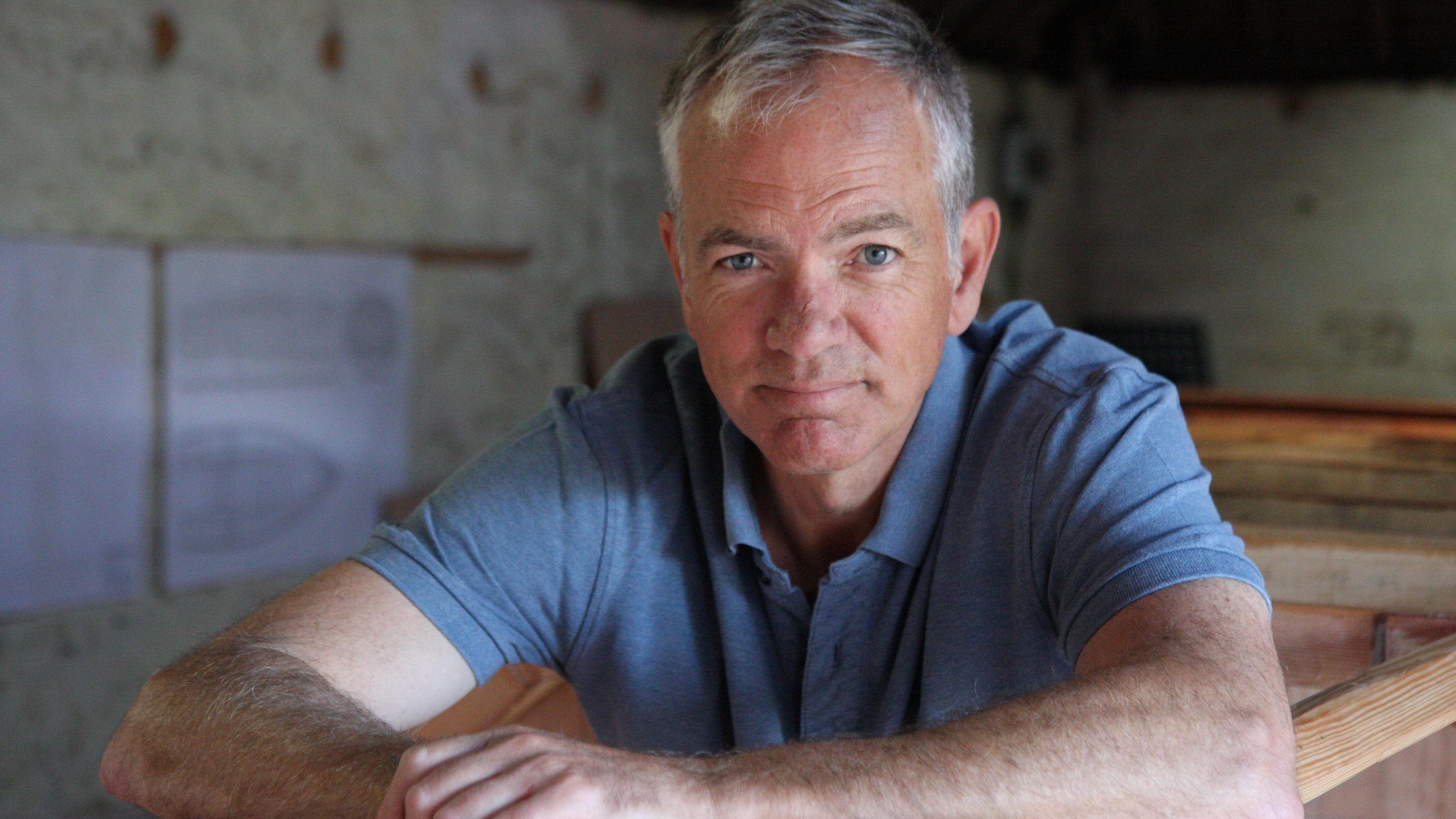 Adam Waugh in the shed where he built his boat, looking into the camera. He has short white hair and blue eyes. He is wearing a blue polo shirt.