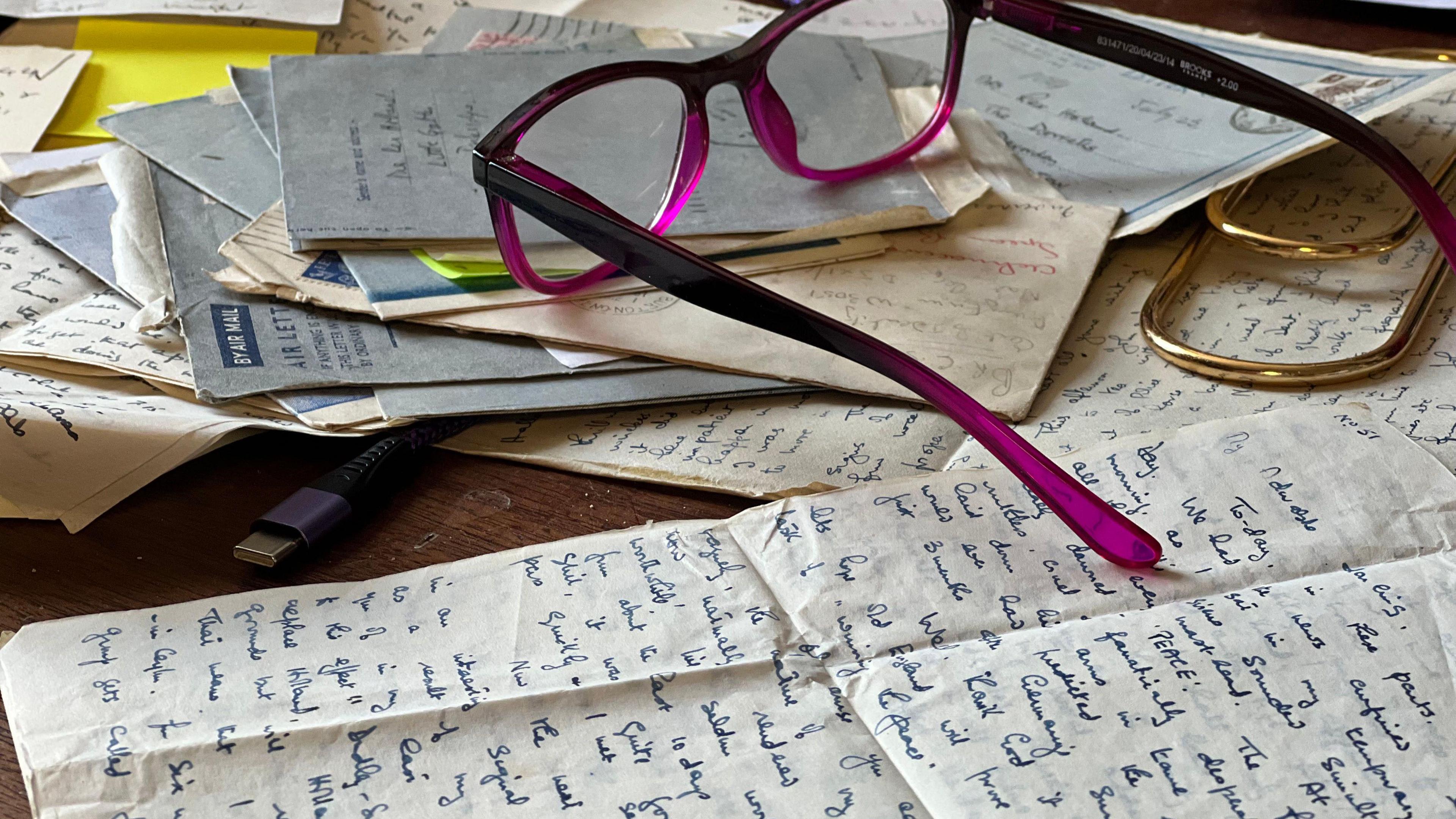 A pile of old handwritten letters and envelopes with a pair of pink framed reading glasses on top of them. 