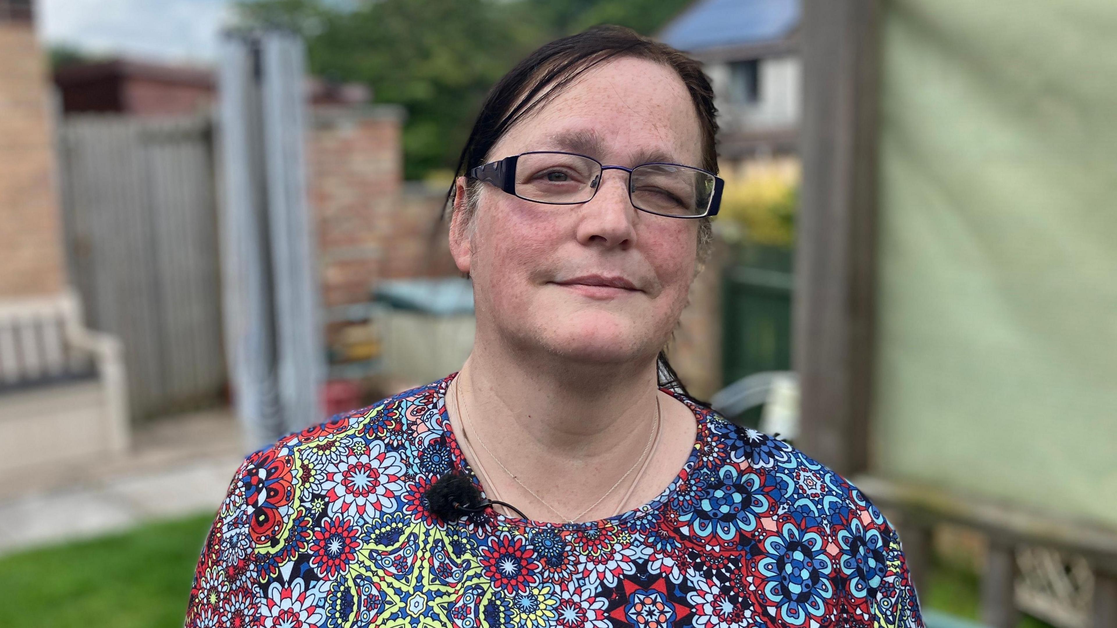 Louise Birch, a middle-aged woman with dark brown hair, and slight smile, wearing black rectangle glasses thin rimmed with two thick handles, with her left eye closed, wearing a patterned circle shirt, blurred background of a garden, 