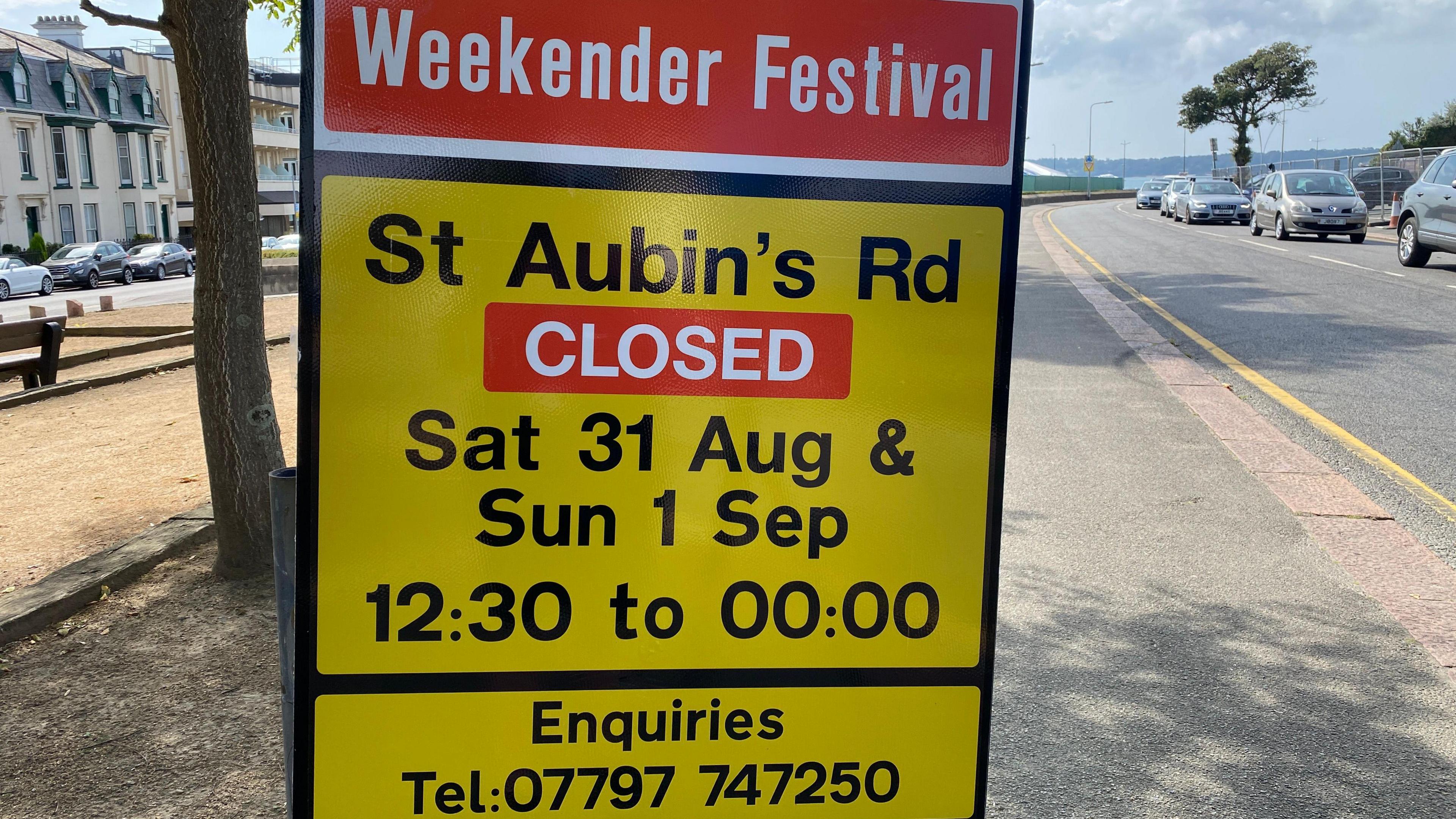 A yellow sign with a red block at the top saying 'Weekender Festival' and details of the road closure. Behind is a walkway, a road on either side with queues of cars, on a grey cloudy day.