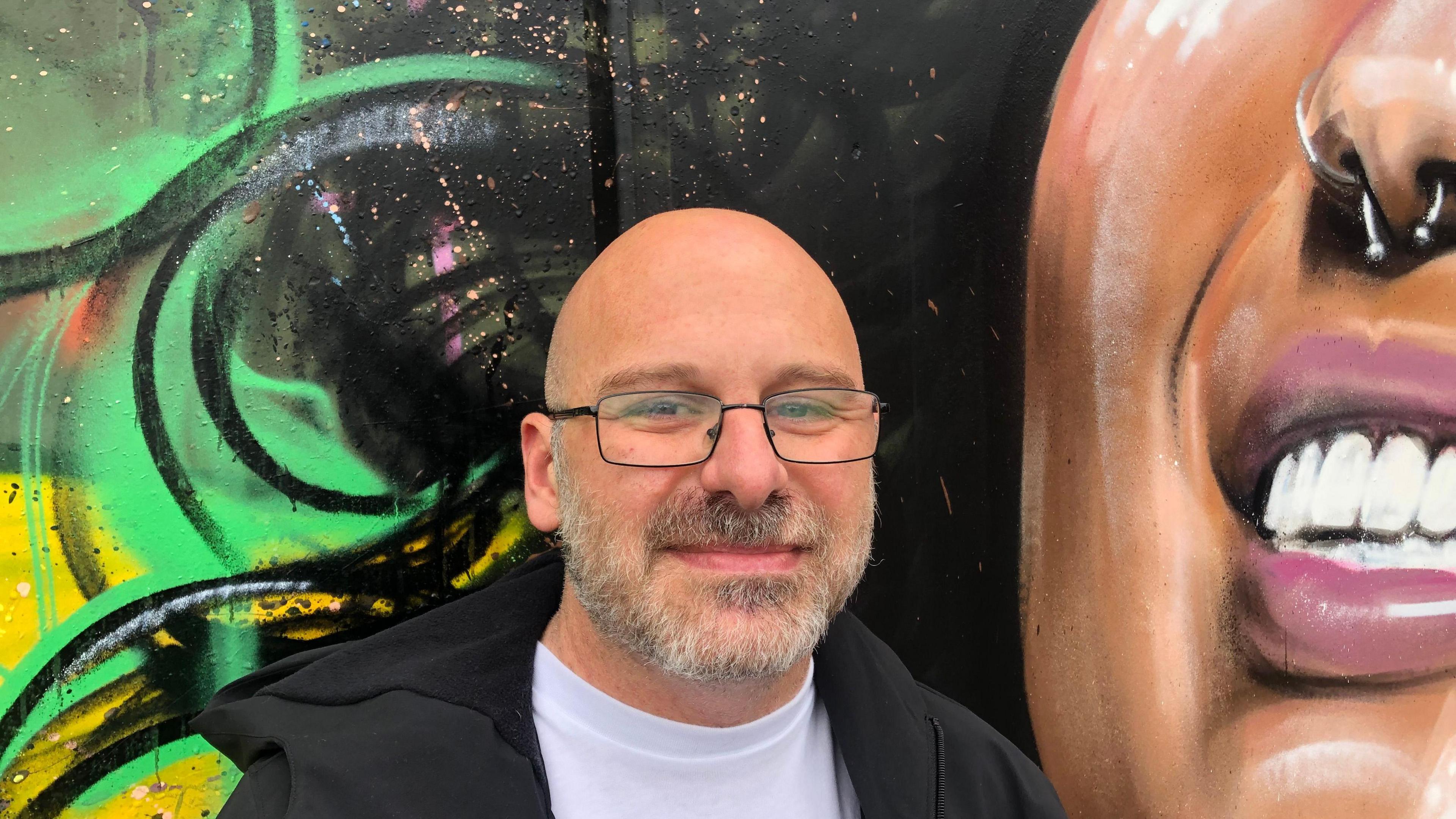 Dave Farrington, 41wears a brown/grey beard and moustache with light framed glasses, a black jacket over a white T-shirt. He is standing in front of part of the mural featuring the smiling woman's lower face.