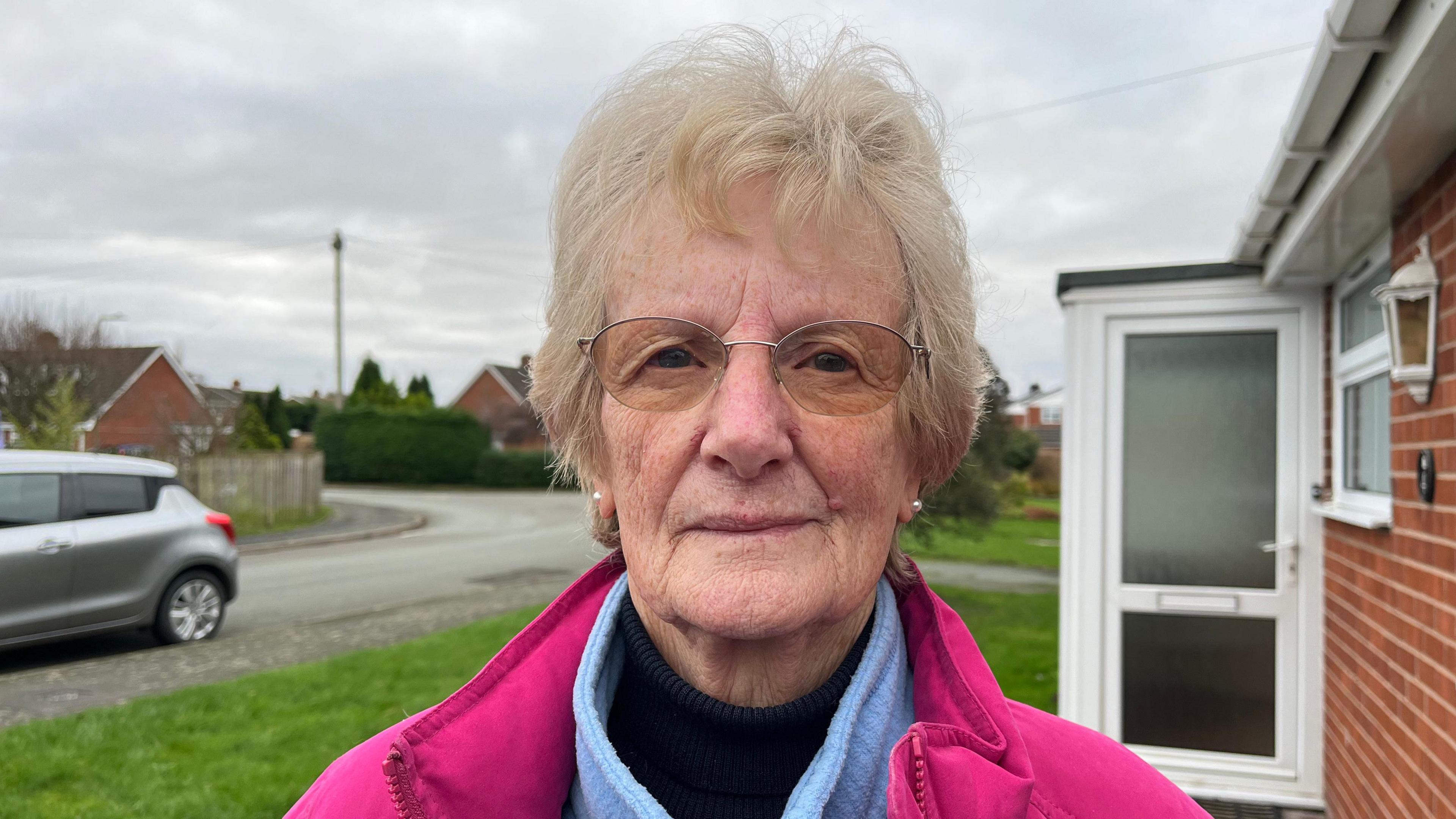 Maureen looking down the camera, she's wearing a pink winter coat with a blue fleece and a navy rollneck top underneath. Behind her you can see Pinewood Road in Shawbury with rooftops and cars visible, the sky is grey and overcast. 