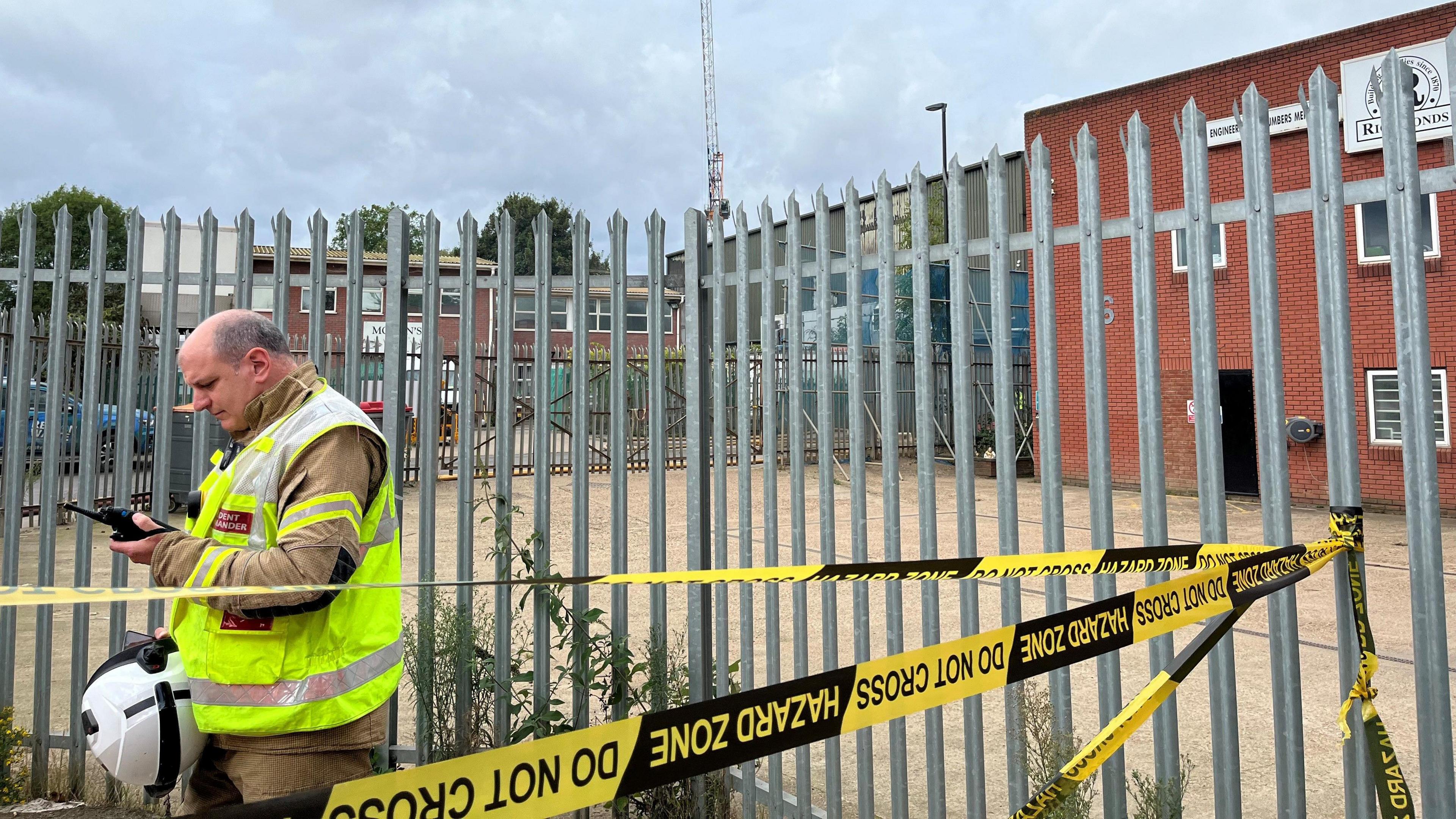 Firefighter stands in front of metal-barred fence which has a yellow and black hazard tape wrapped around it looks down at a controller in his hand 