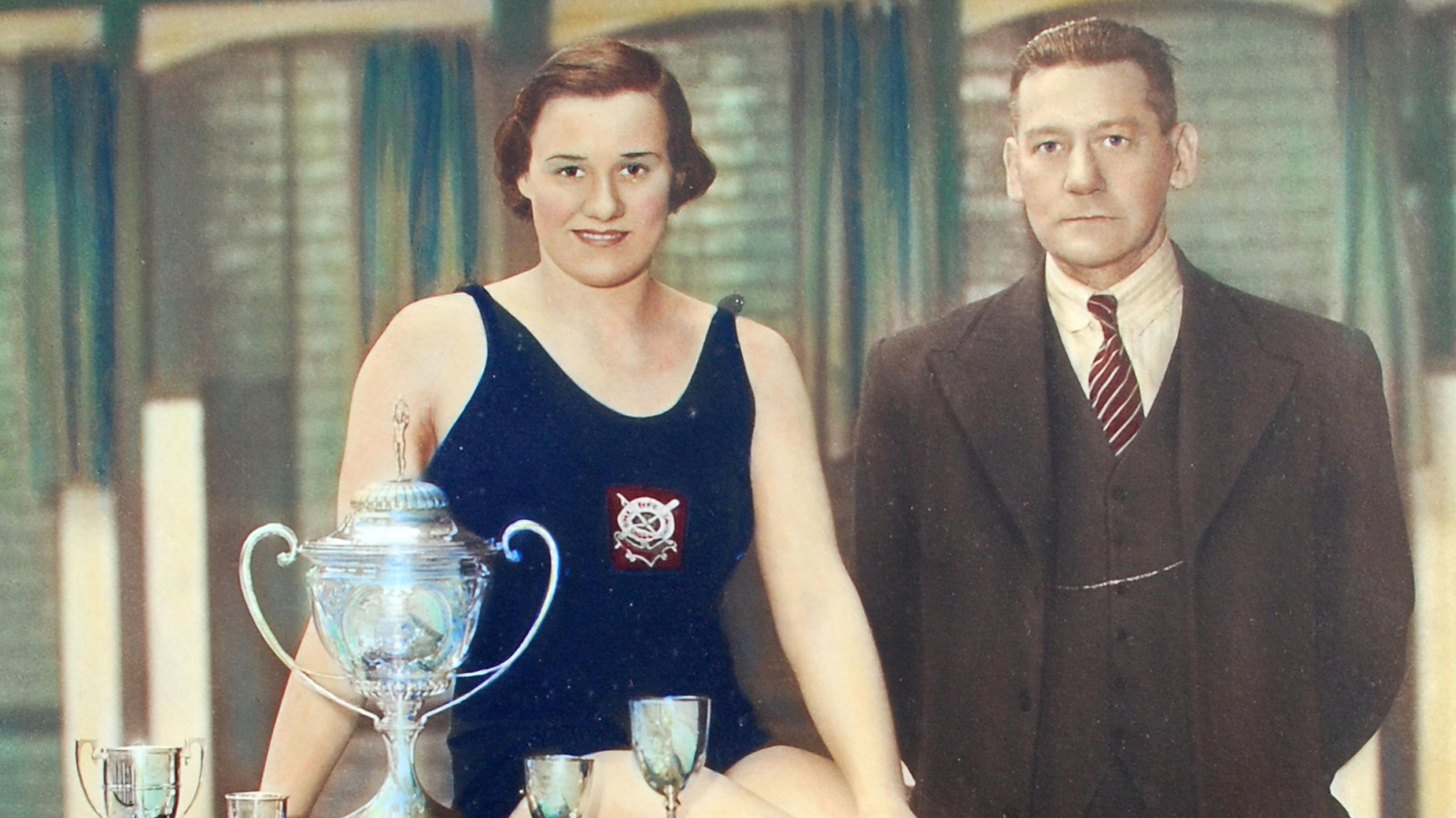 A sepia-toned image of a female swimmer wearing a Navy blue swimming costume sitting behind four trophies accompanied by a man in a brown suit.