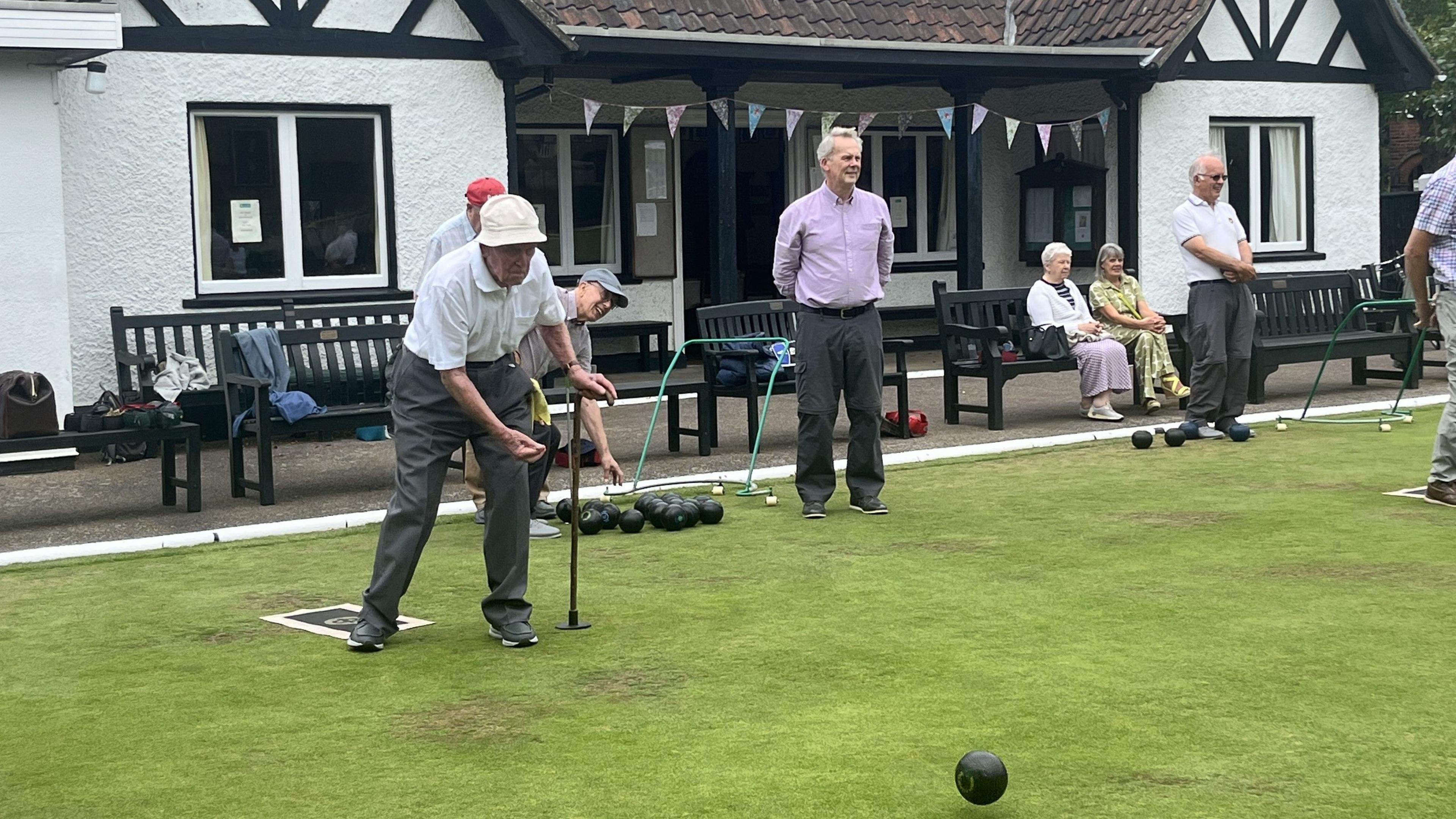 A man playing lawn bowls is wearing a white hat and shirt and grey trousers and trainers as other players watch on from benches in front of a white clubhouse which has black wooden beams on the outside.