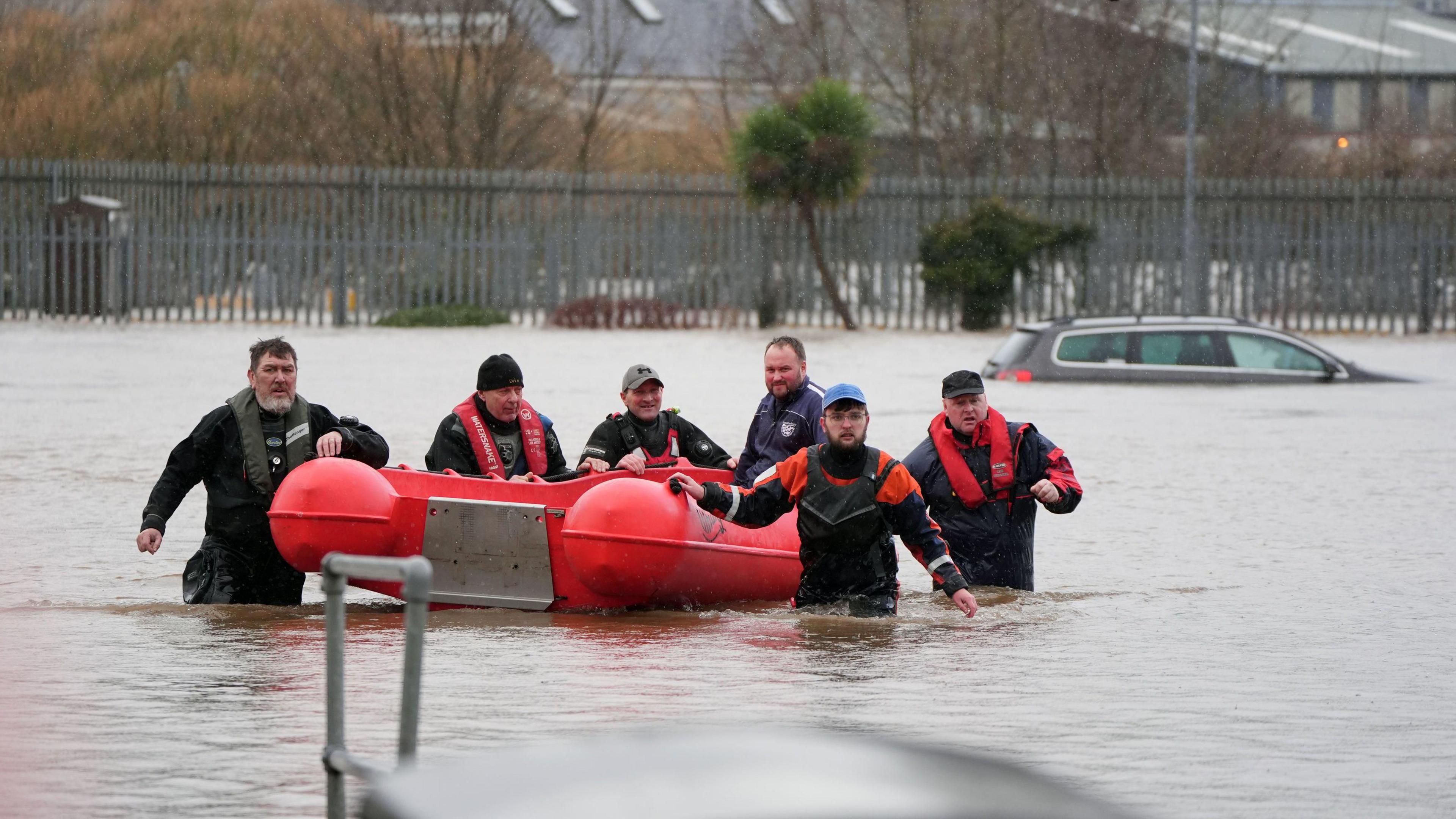 Five men in waterproof waders pull a red lifeboat through floodwater. The water covers their entire legs. There is a man sat in the boat. Behind them, half a car pokes out of the water