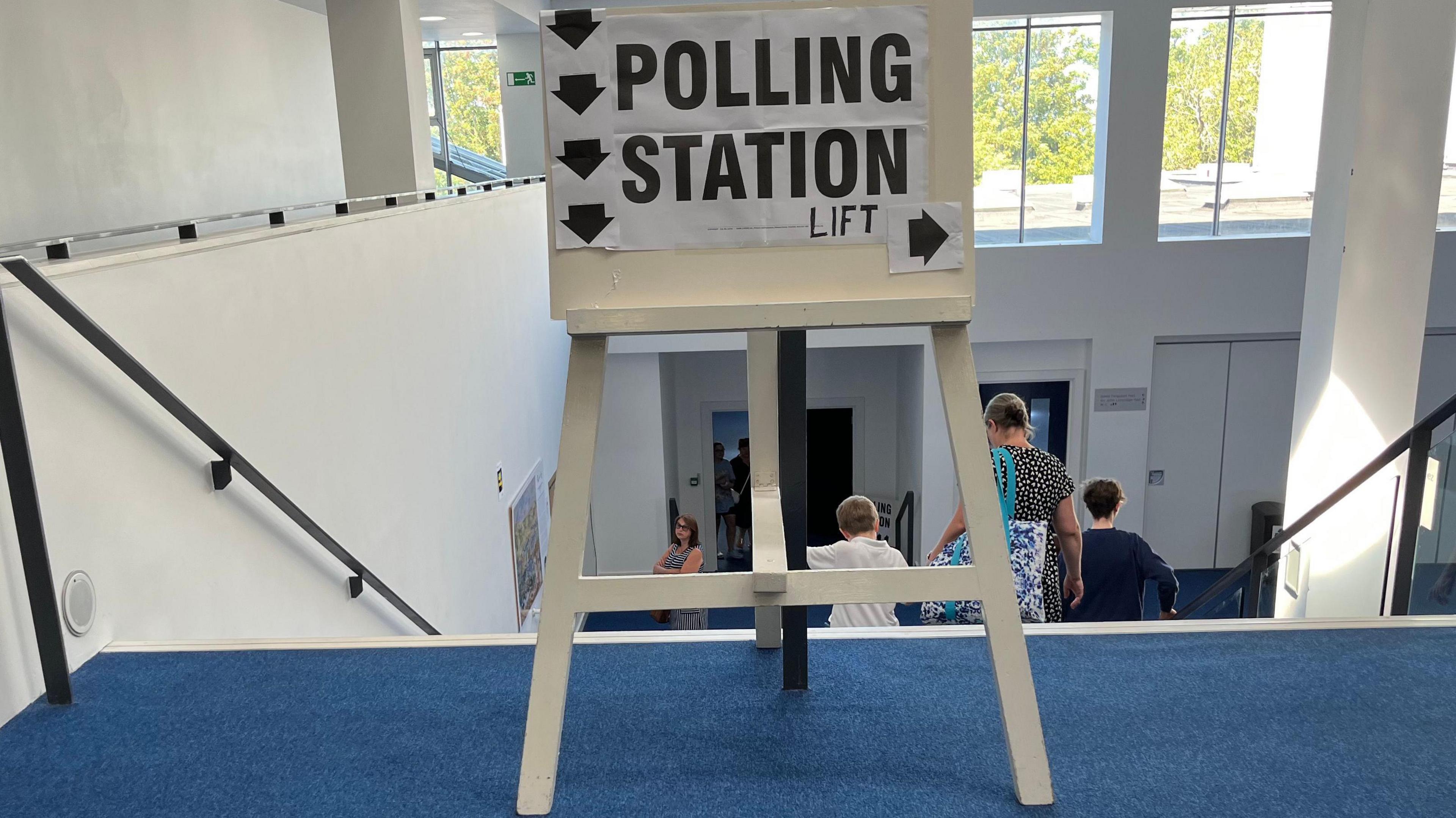 A white wooden sign which says Polling Station with arrows saying down the stairs and a sign to the right indicating the location of the lift.
The sign is at Beau Sejour in Guernsey.
