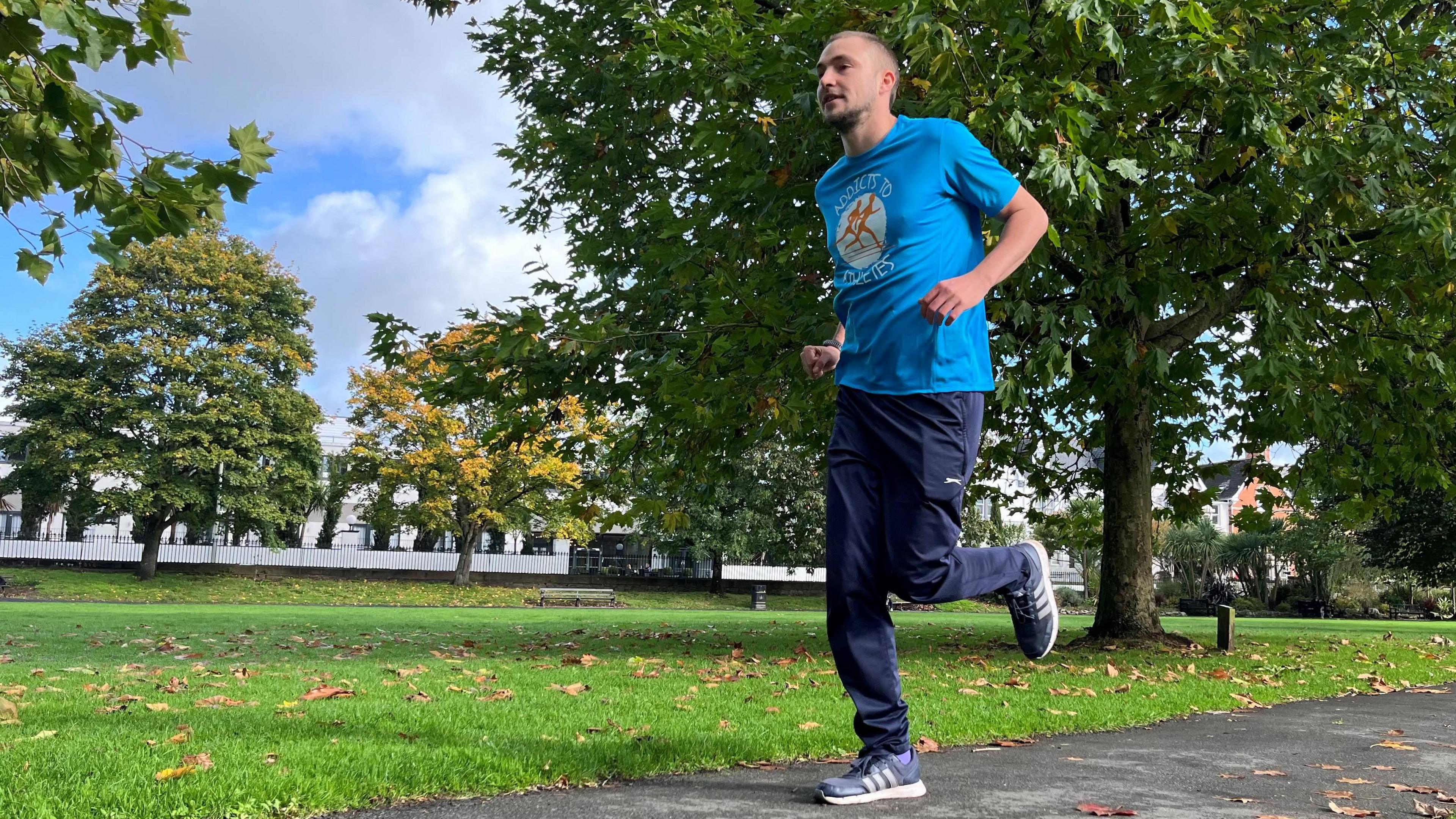 Connor Shannon wearing a light blue T-shirt and dark blue running trousers and trainers. He is running through a park in Barnstaple.