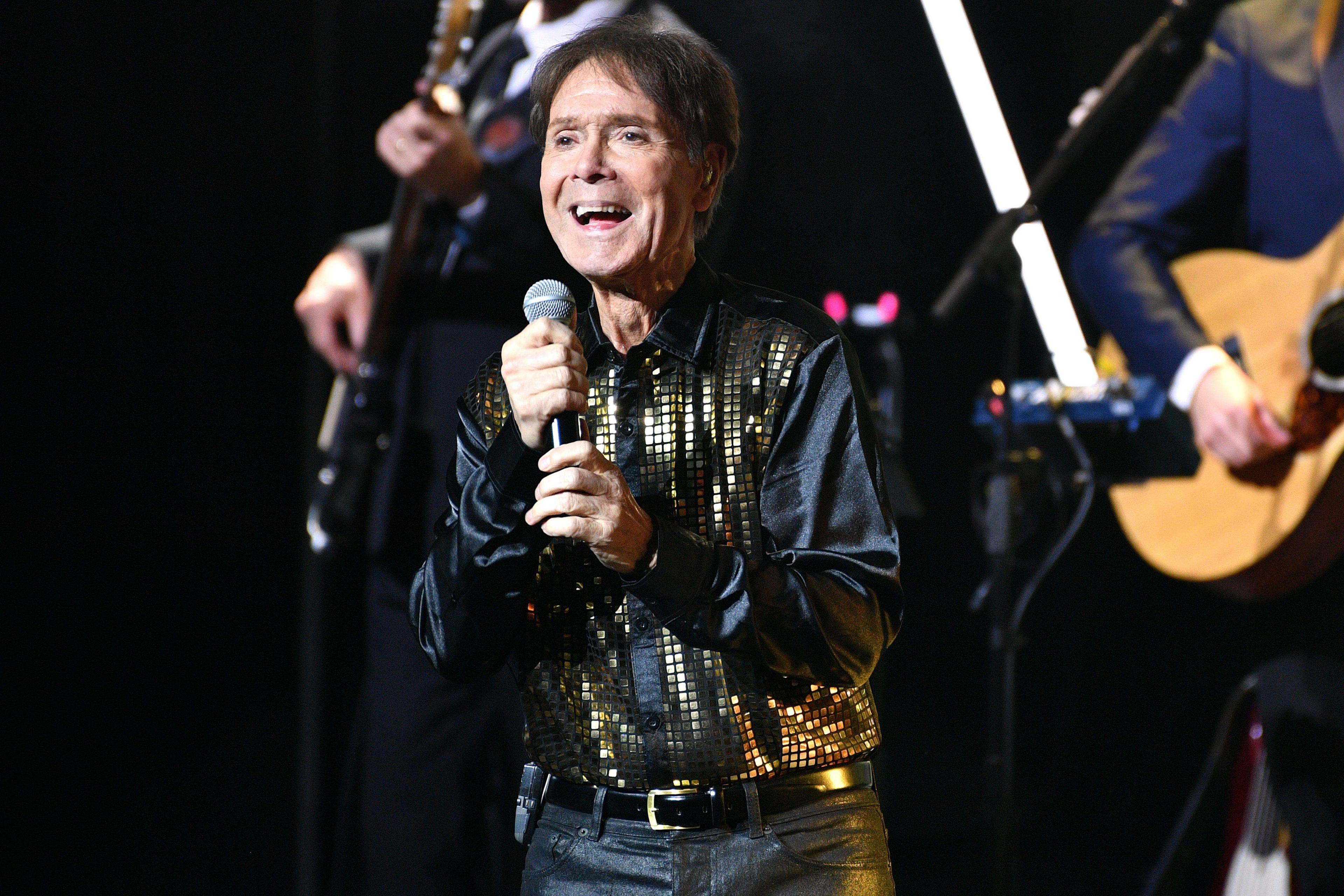 Sir Cliff Richard on stage at a concert holding a microphone and smiling. He wears a black jacket with gold sequins and dark shiny trousers. Two guitarists are behind him.
