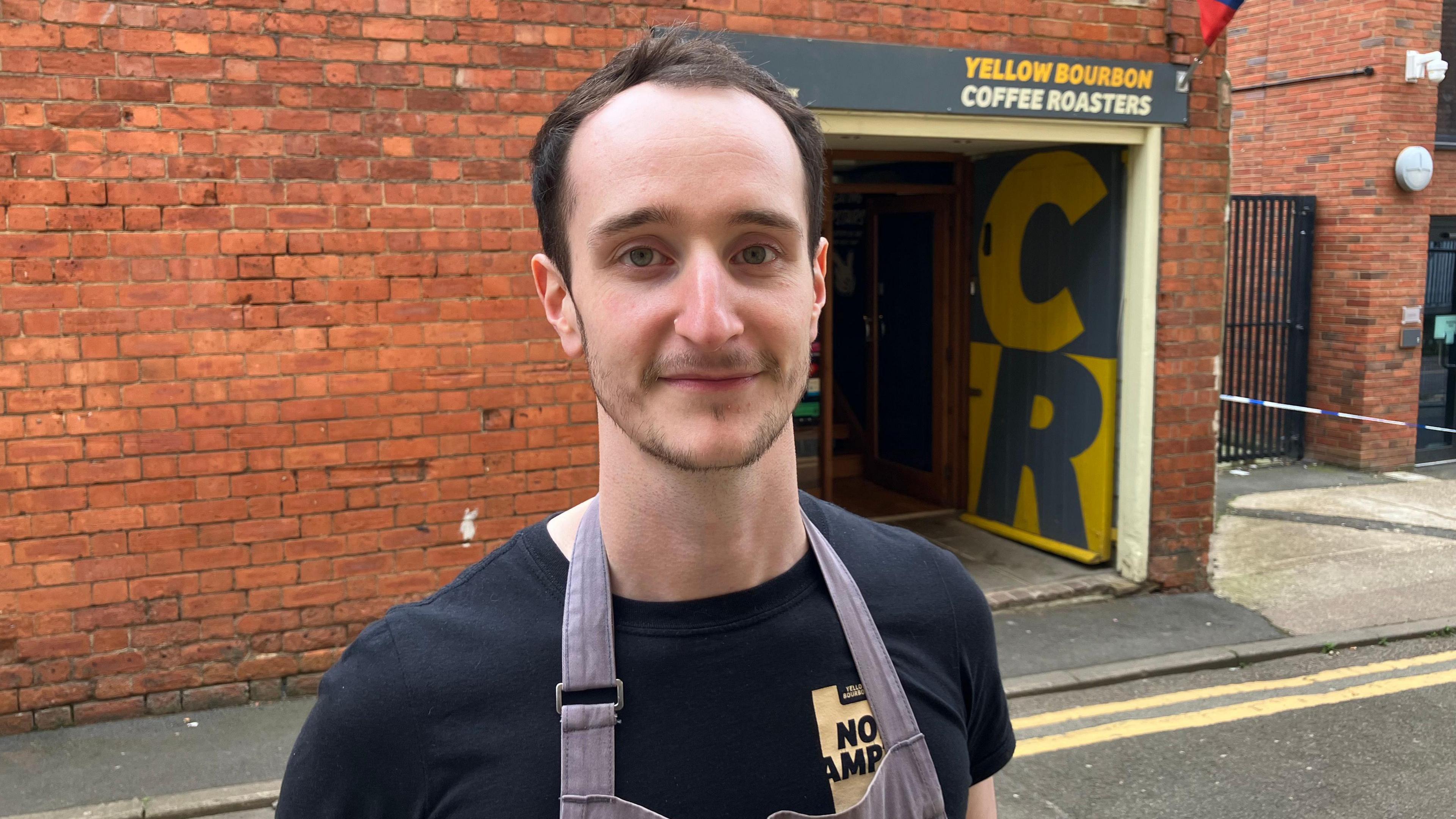 Sam Peace is wearing an apron and a black t-shirt and standing in front of the shop 