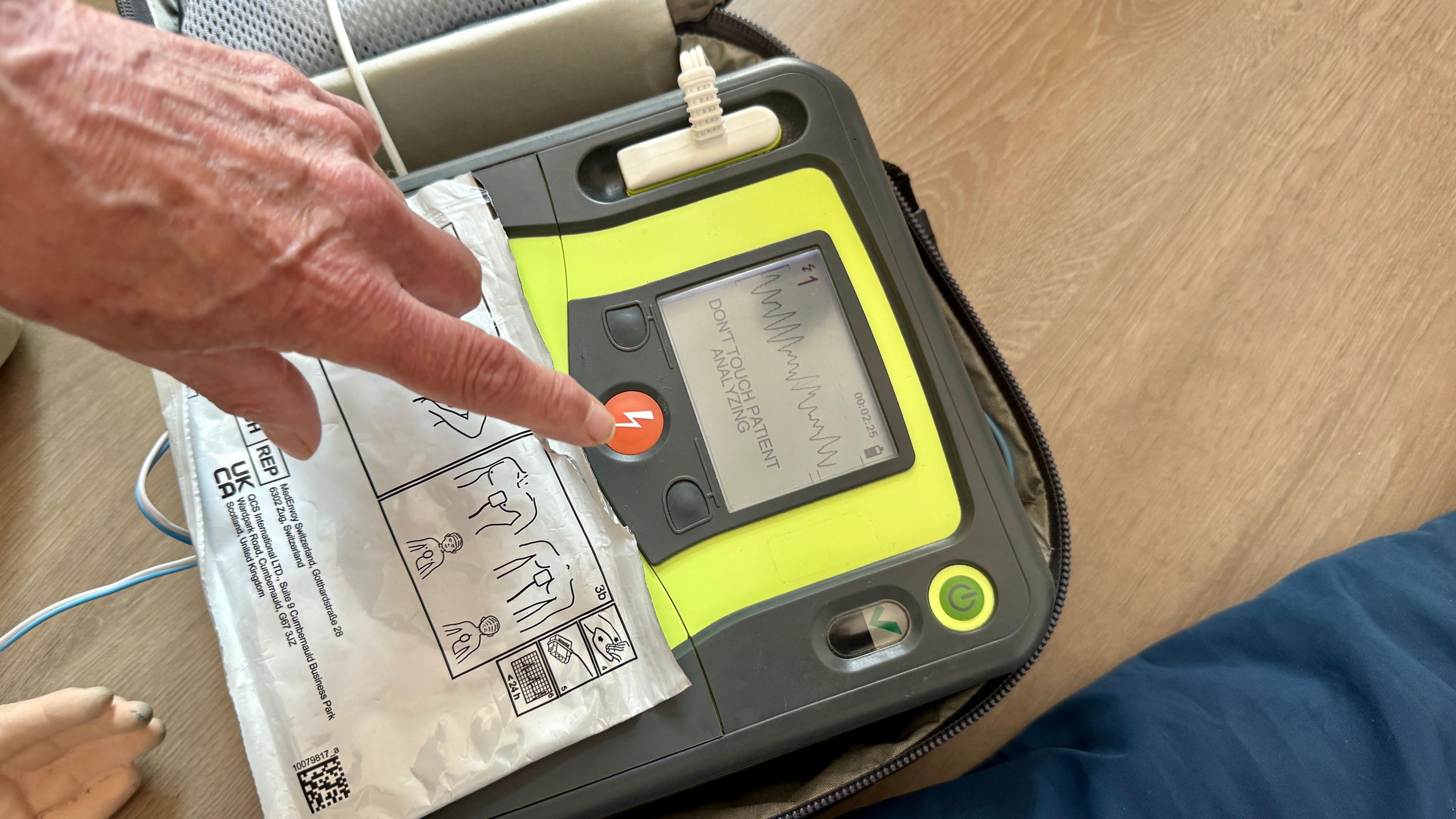 A defibrillator on the floor being used. It has a fluorescent yellow and black casing and a finger is floating over the power button. You can't see the person inthe picture. There are instructions on a large white sticker over the front of the device.