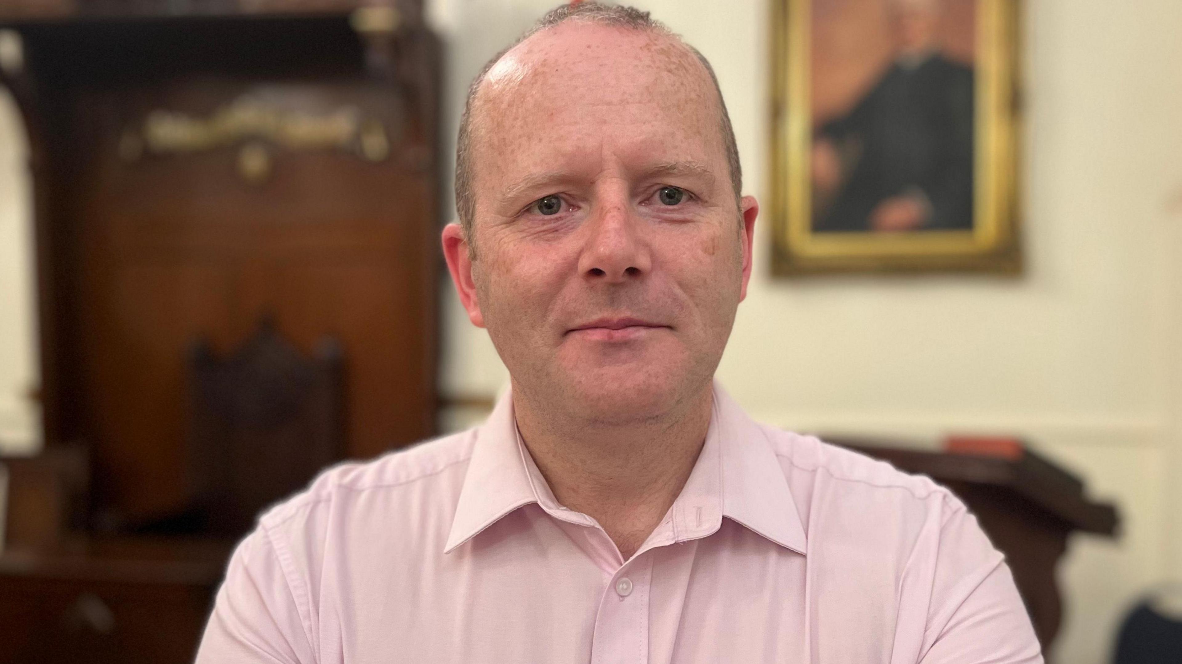 Ron McDowell in a pink shirt. There is a photo on the wall behind him.