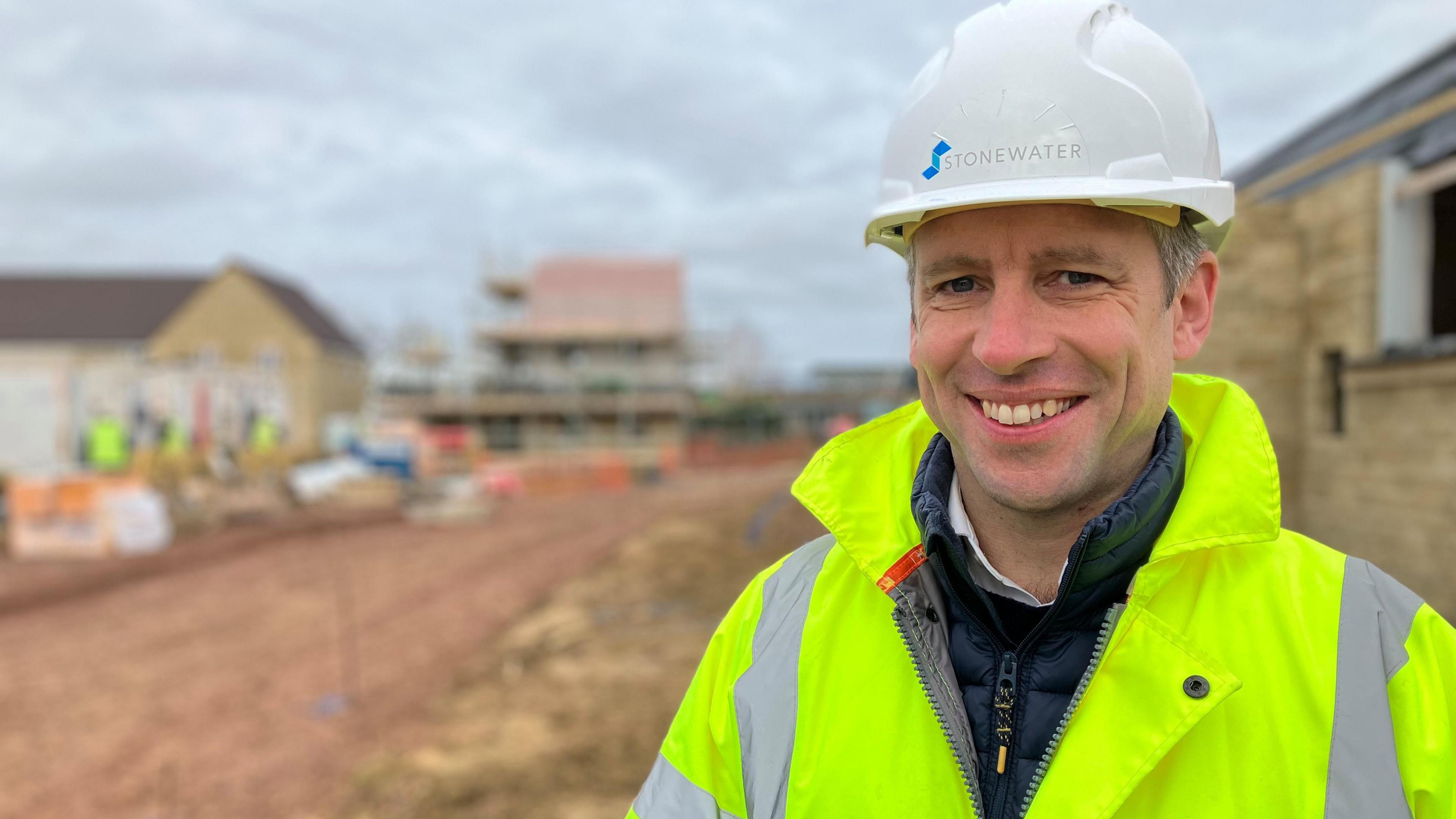 Jonathan Layzell from Stonewater wearing a hard hat and high visibility jacket on a building site at Martock in Somerset. He is smiling at the camera and new homes in progress can be seen in the background