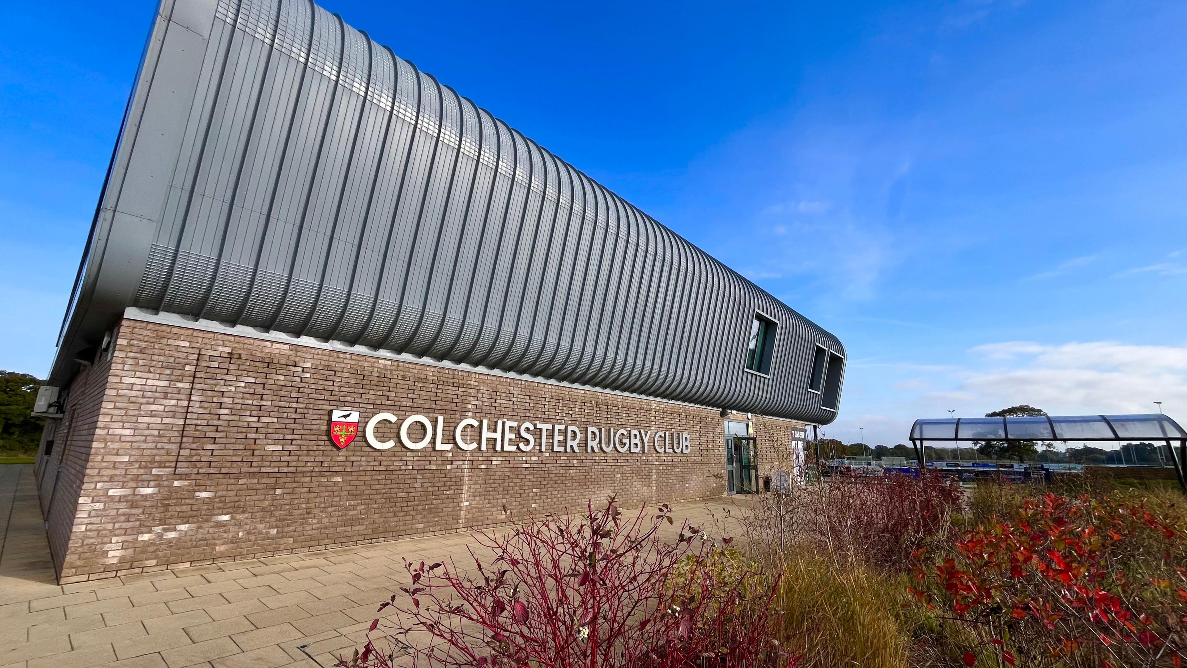 The outside of Colchester Rugby Club's clubhouse. It is a brick building with the club's logo on the exterior and bushes in front of it.