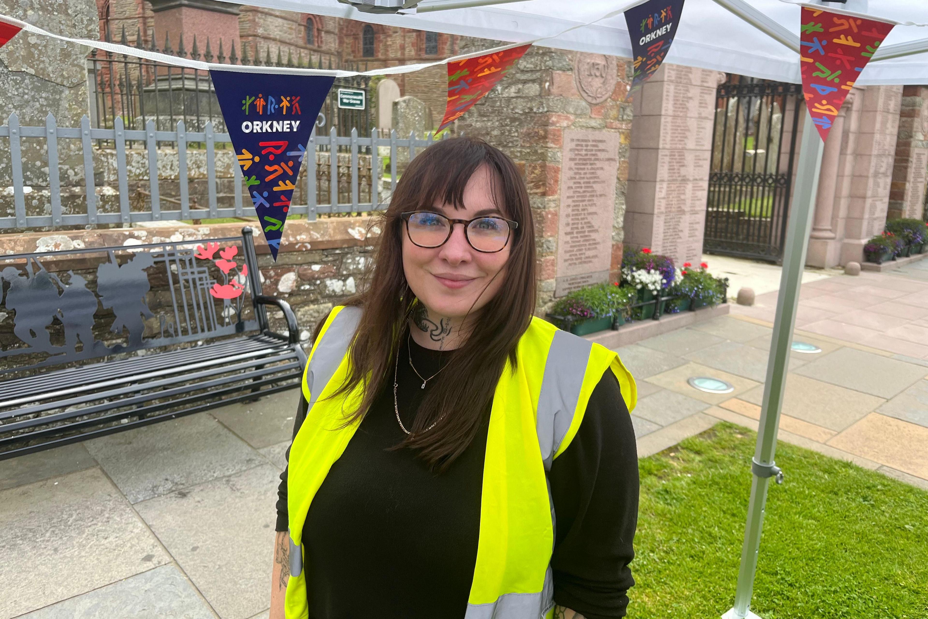 A woman in a high-viz vest with glasses on and blue and red bunting hanging from a marquee in the background.