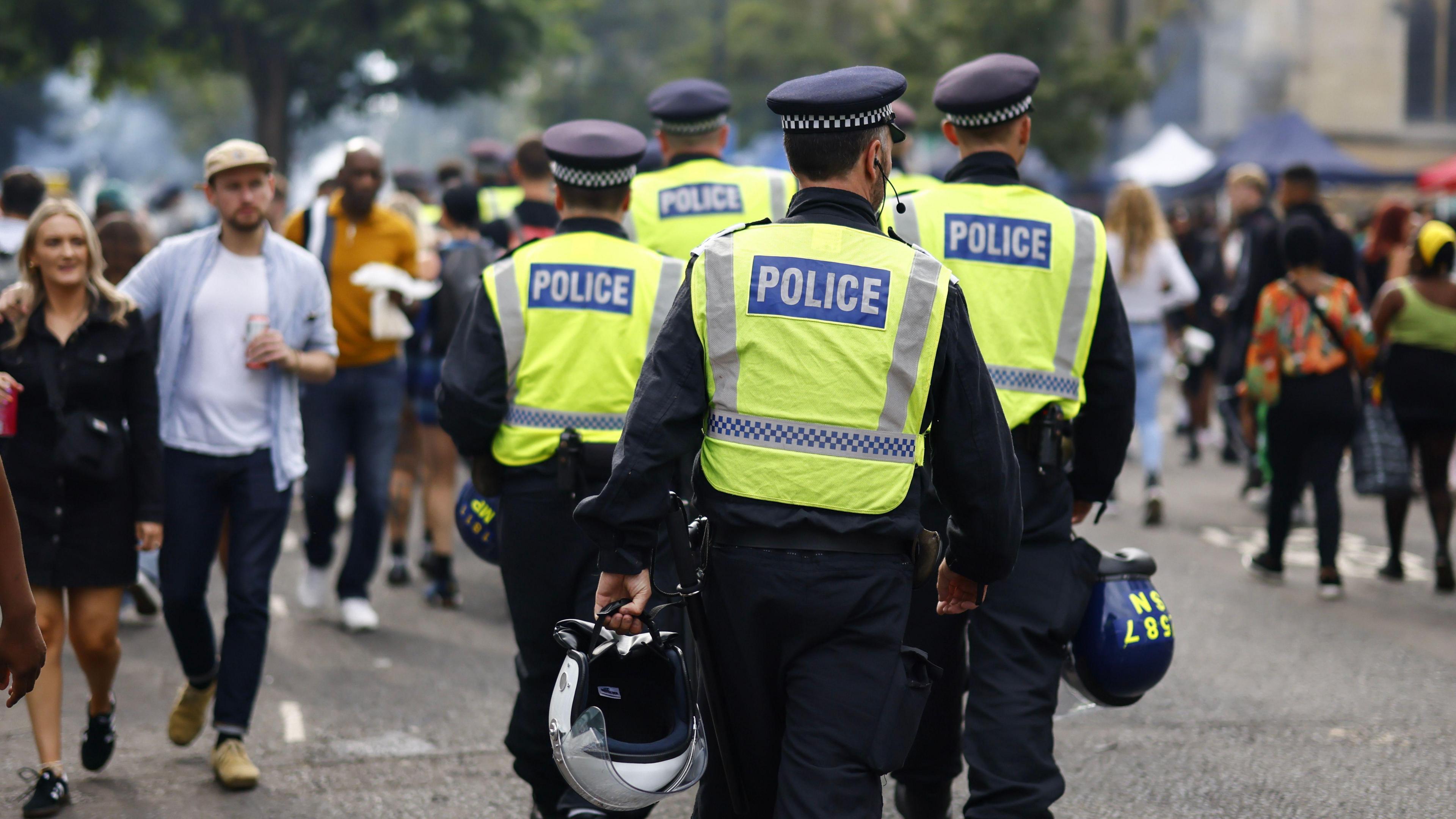 The backs of five police officers on patrol during the first day of the Notting Hill Carnival in London in 2023. People are seen walking on the street. The police officers are carrying helmets.