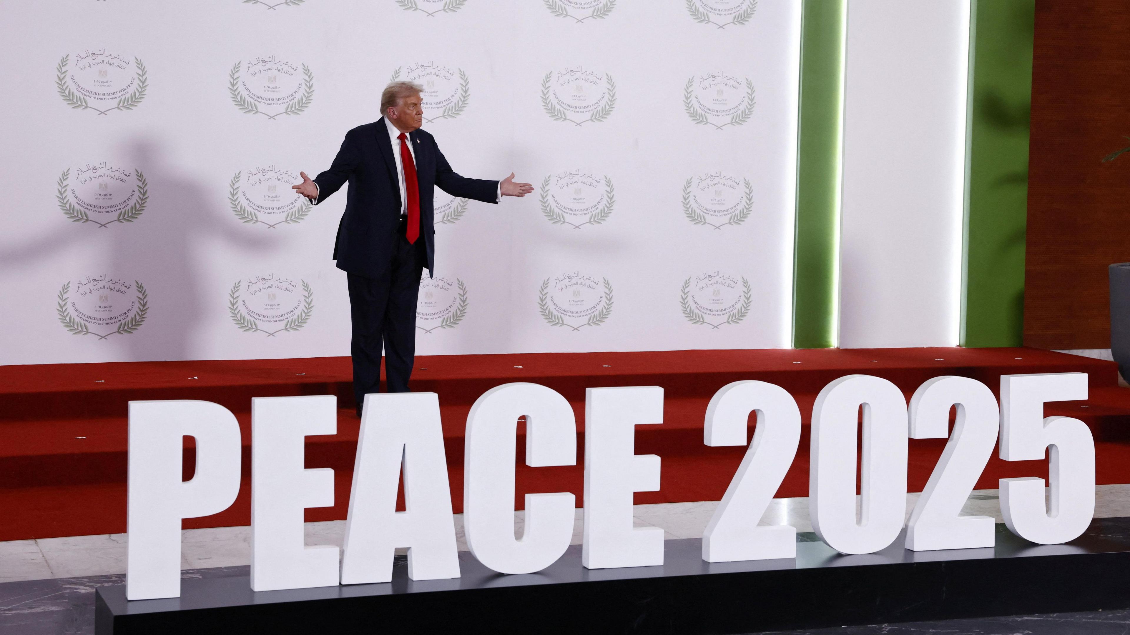 President Trump standing on stage with his arms outstretched behind a large sign which says 'Peace 2025'. 