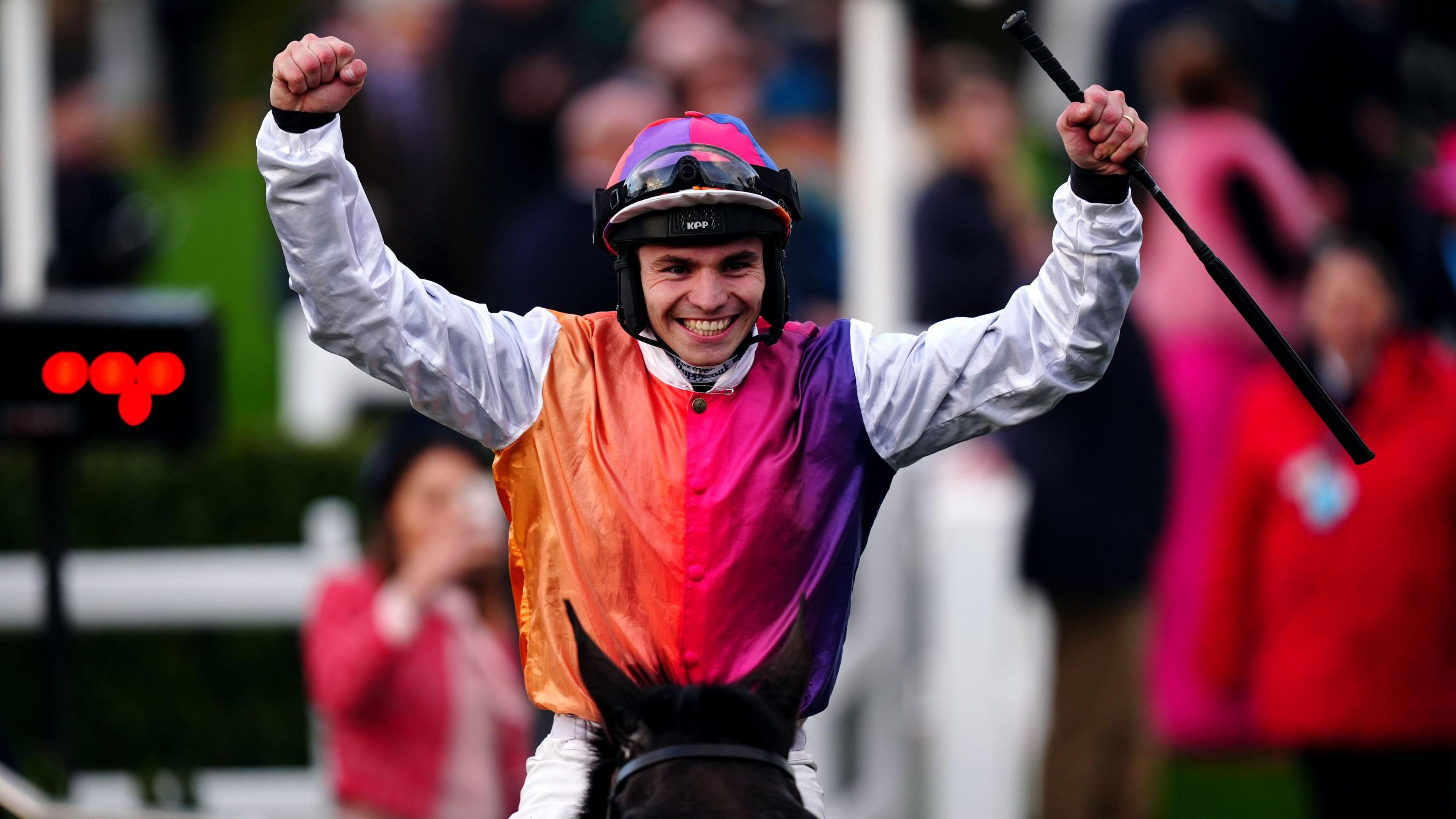 Ben Jones raises both arms in the air in victory while he rides his horse. He is wearing a racing shirt with red, pink and purple colours and white long sleeves. He has a purple and pink helmet.