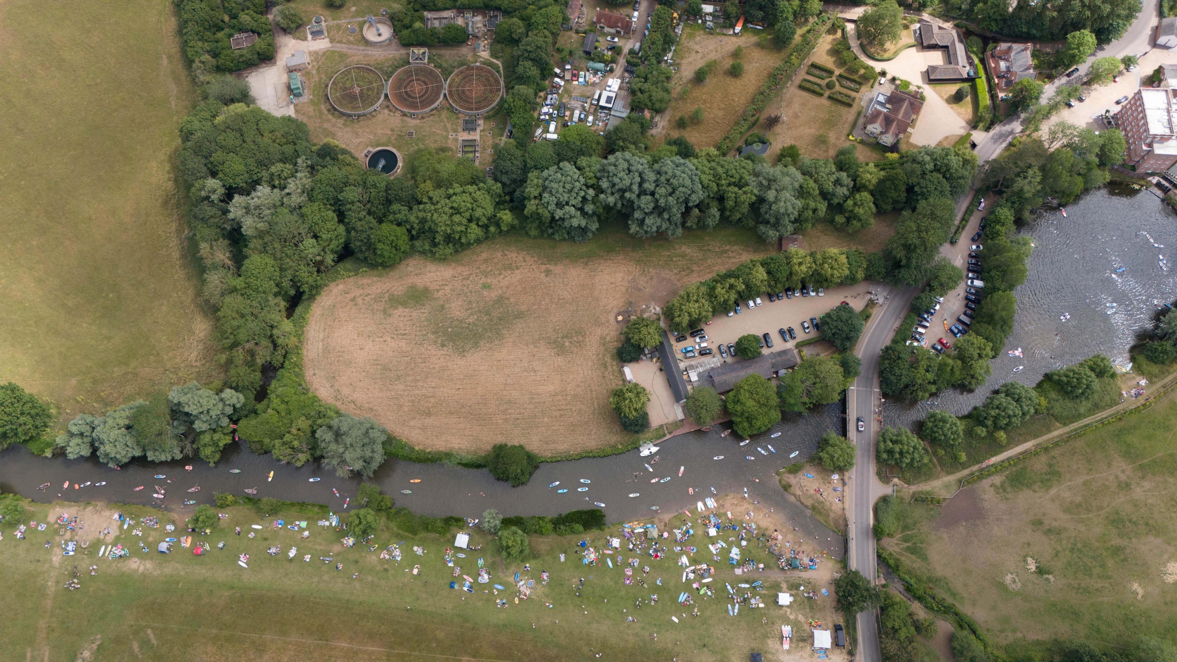 An aerial shot of a river, showing large amounts of people on the river and on one side of it, also a sewage treatment works to the top of the image.