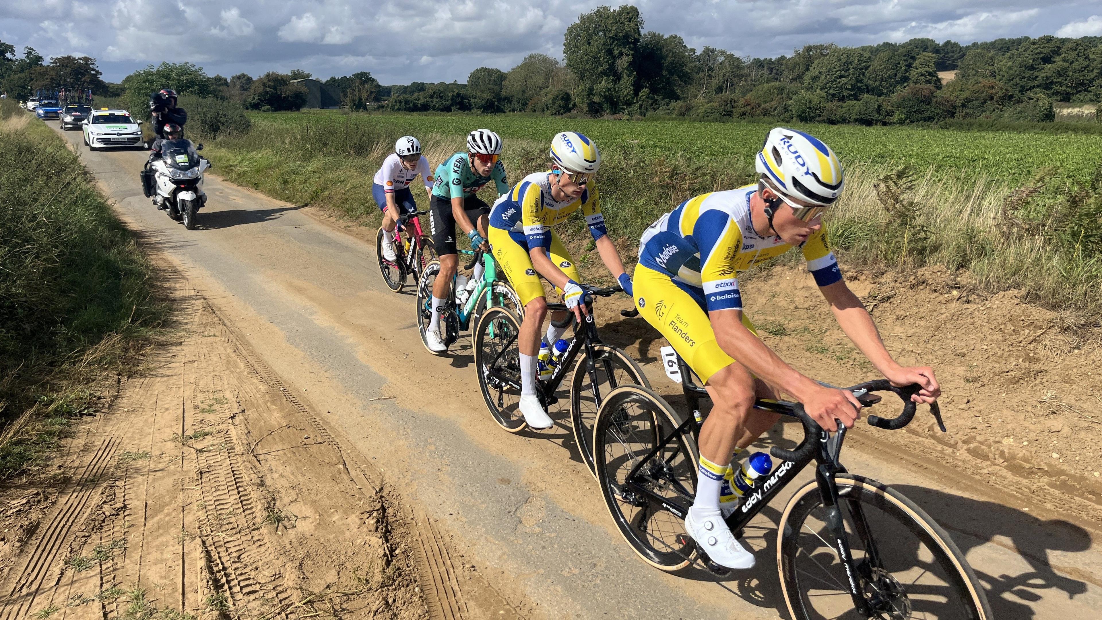 Cyclists race along a single track country road. They are followed by race support vehicles. 