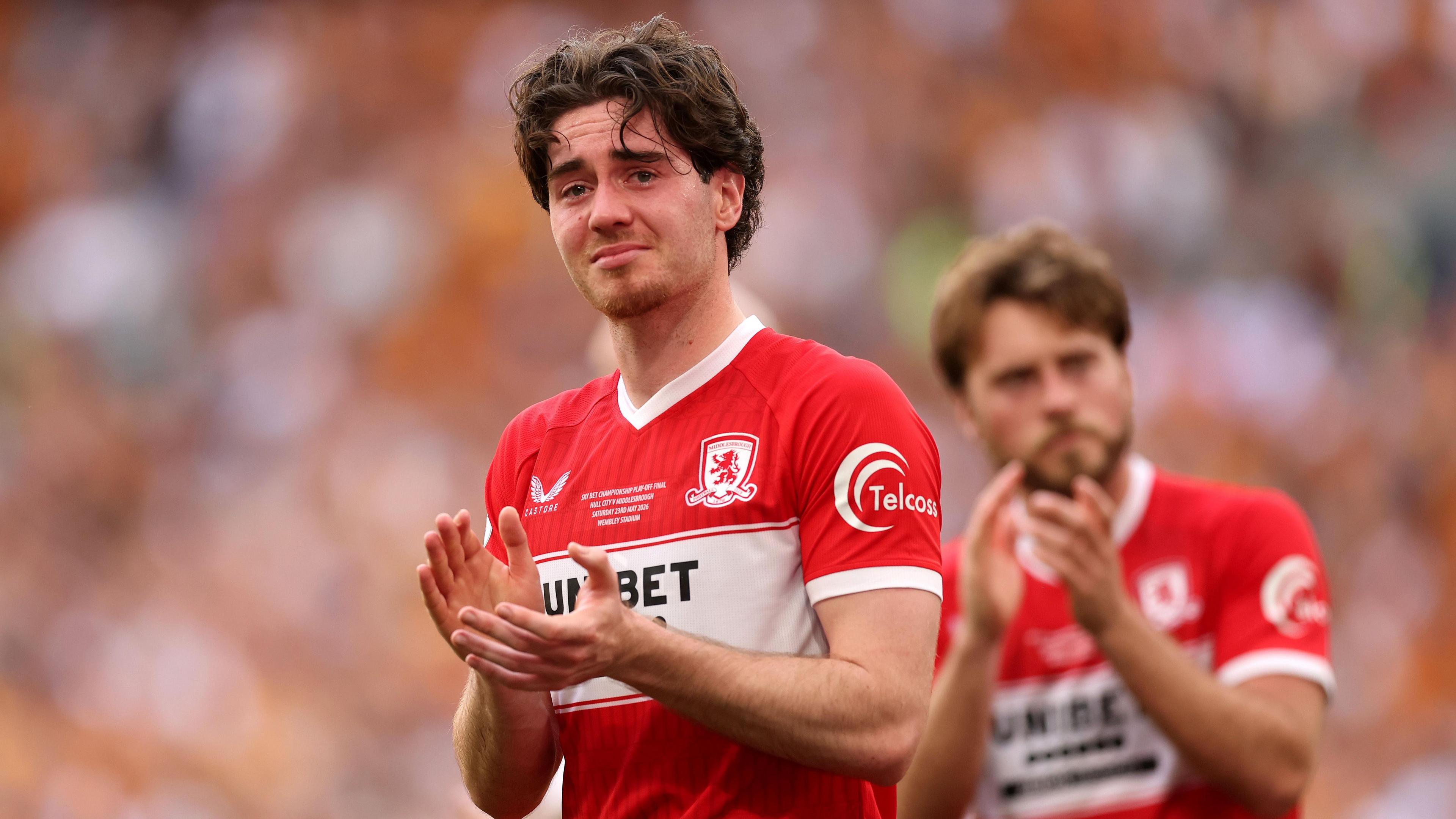 A photo of Hayden Hackney, wearing Middlesbrough's red and white home kit, applauding the fans while looking emotional after their play-off final loss to Hull City in May 2026