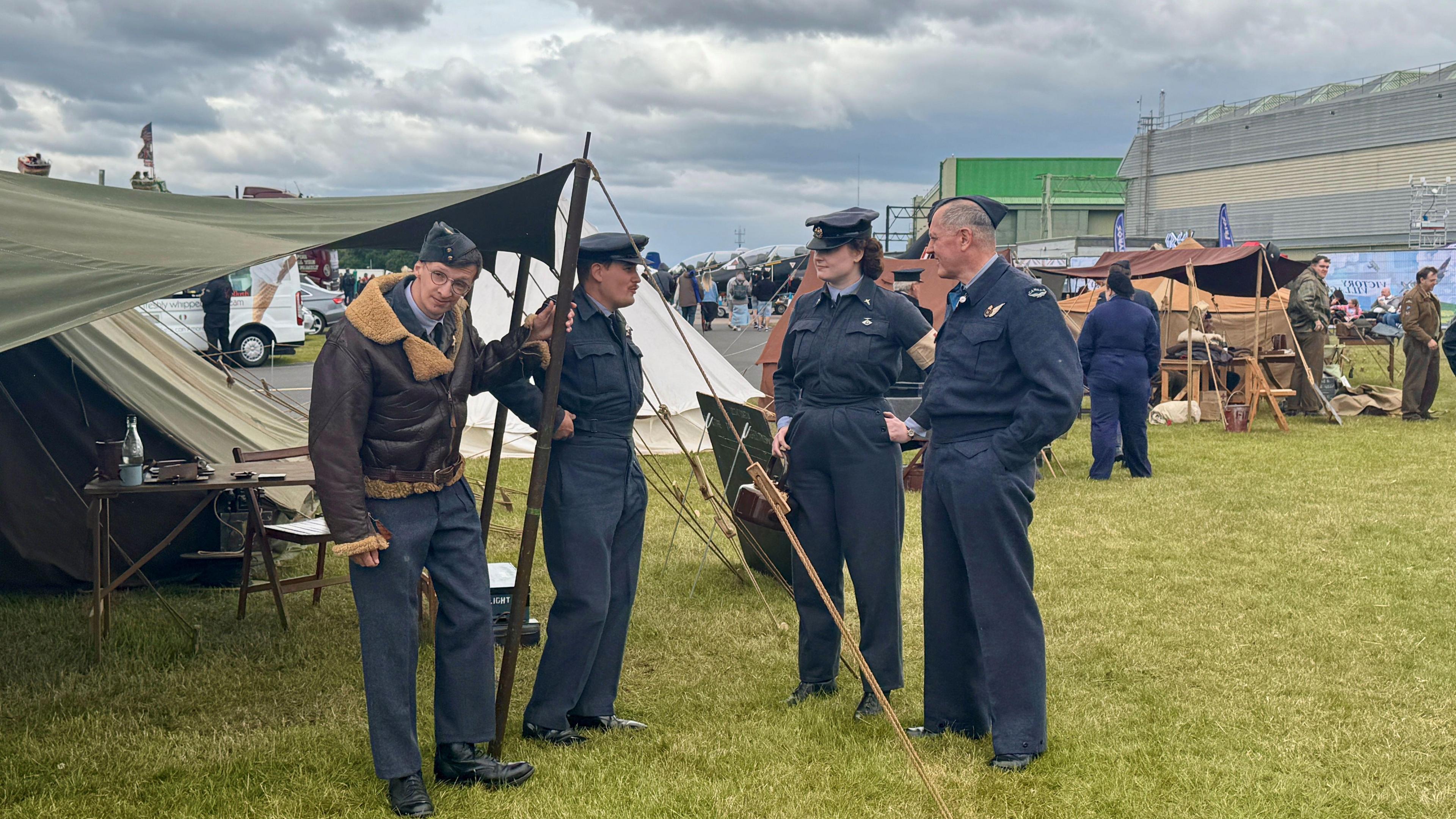 Three men and a woman, wearing air cadet uniforms, with one also wearing a brown bomber jacket, stand talking outside an old-fashioned green tent in the middle of an airfield. More tents and people can be seen behind them.