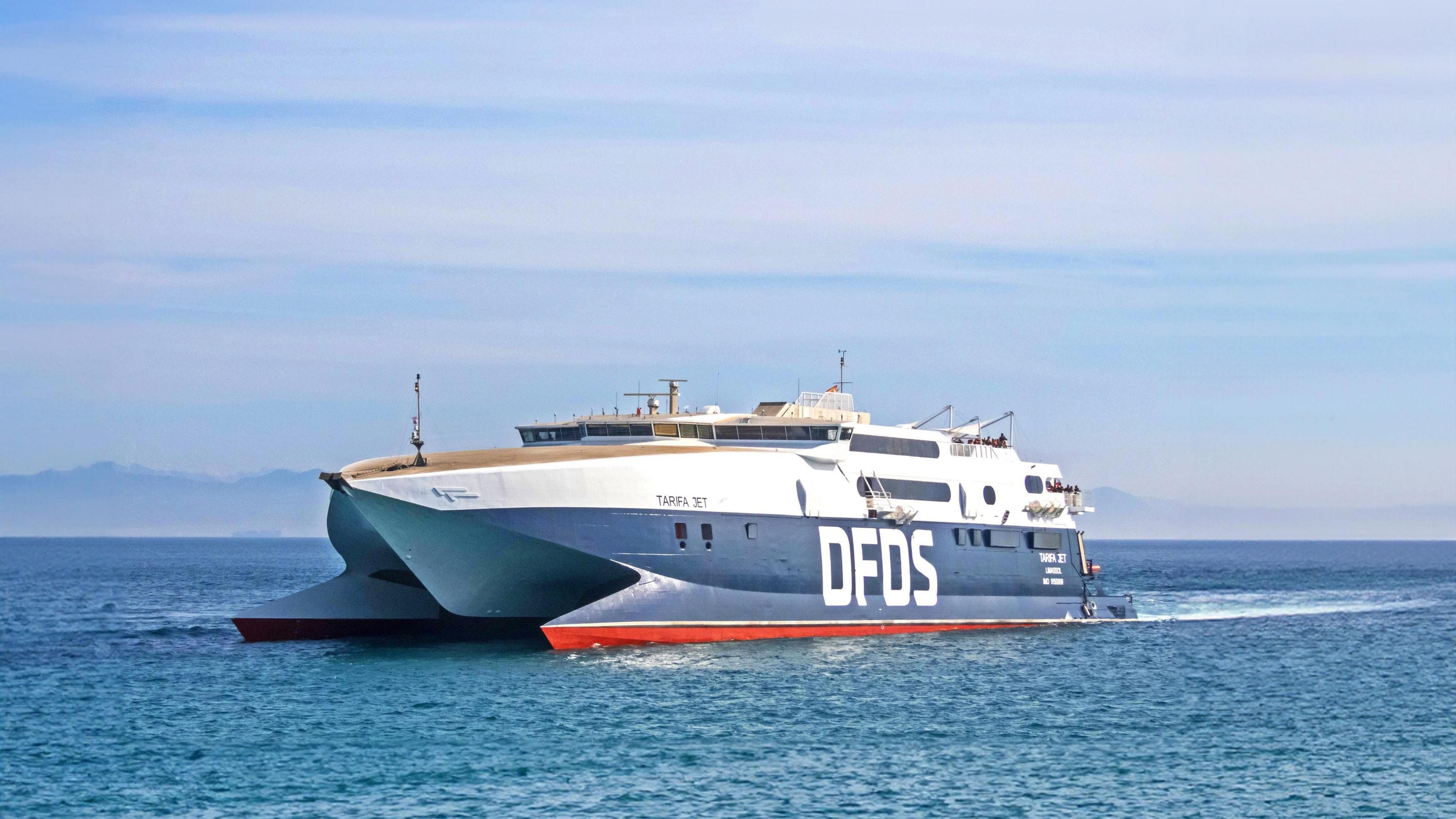 A large ferry sailing across the sea. The ferry has the company name prominently displayed on its side in big white text. The top of the boat is painted in white with the bottom in blue. There is a clear sky with some light clouds.