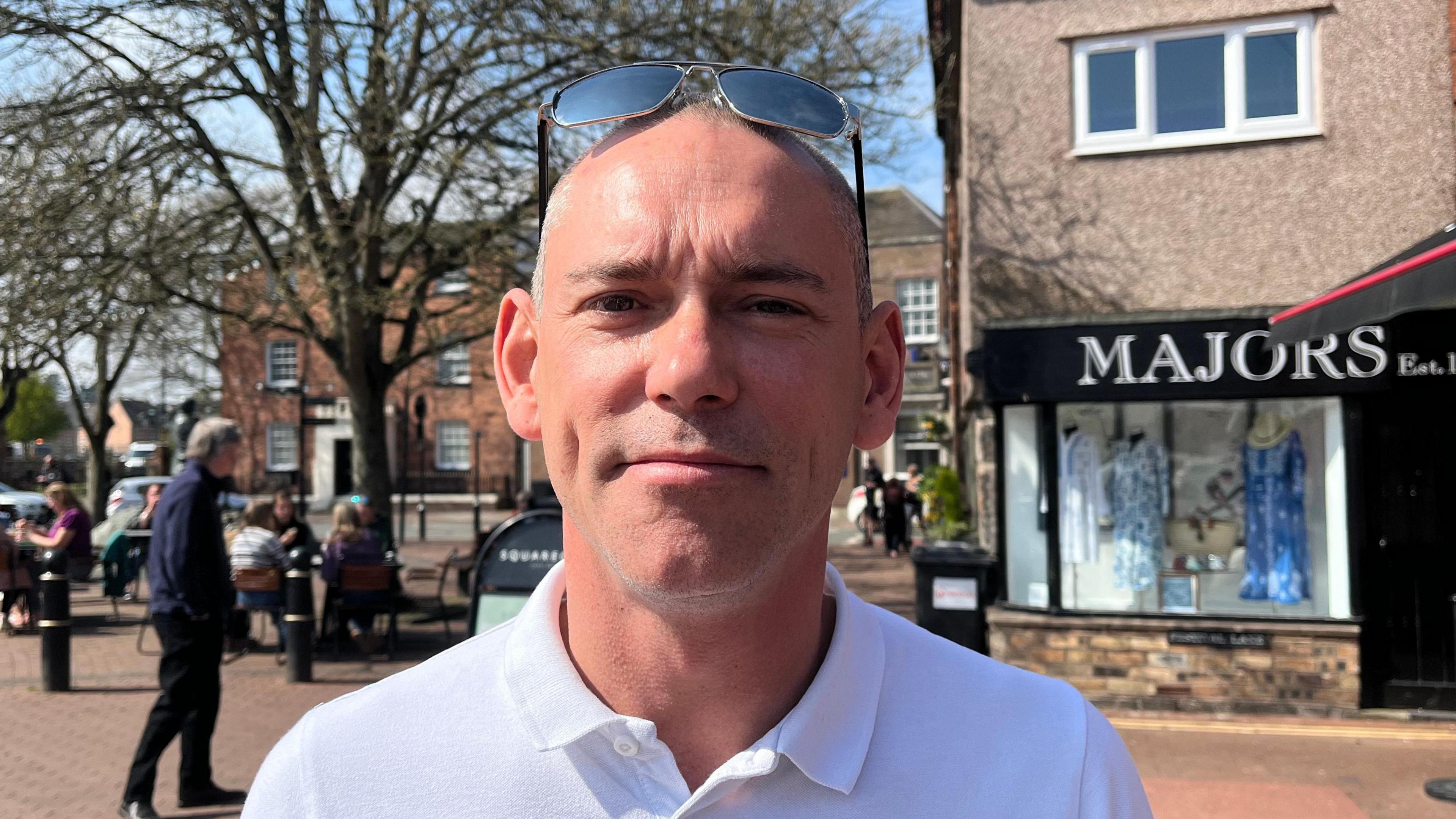 Robert looking into the camera; he's wearing a white polo shirt and has his sunglasses perched on top of his head. He's stood in a town square, with brick buildings beyond his left shoulder and one with a sign reading 'Majors' - a black sign with white writing. There's a large tree over his right shoulder and the sky is blue and the weather is sunny.