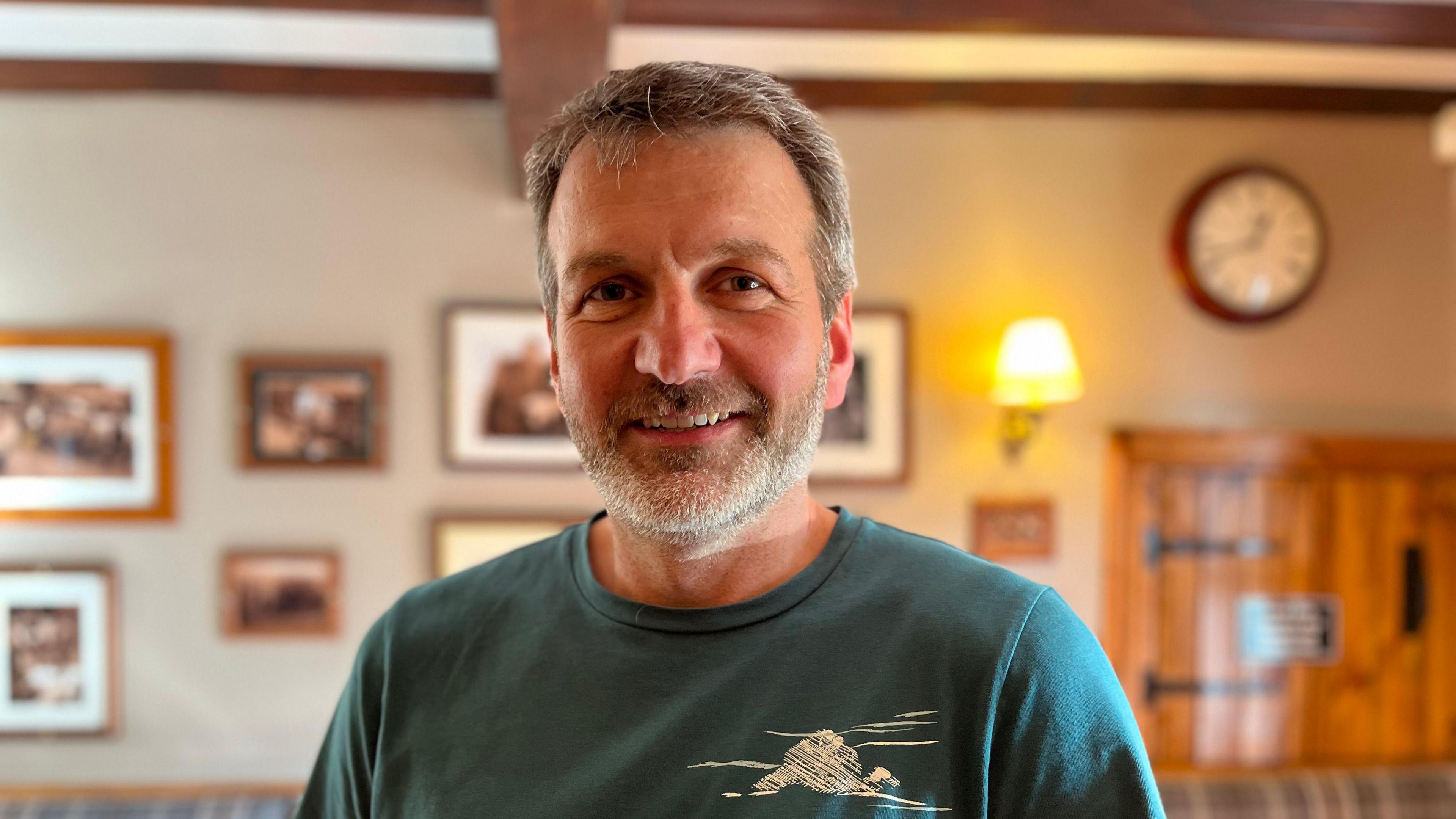 A man with grey hair and a beard, wearing a green top is standing in front of some pictures on a wall.