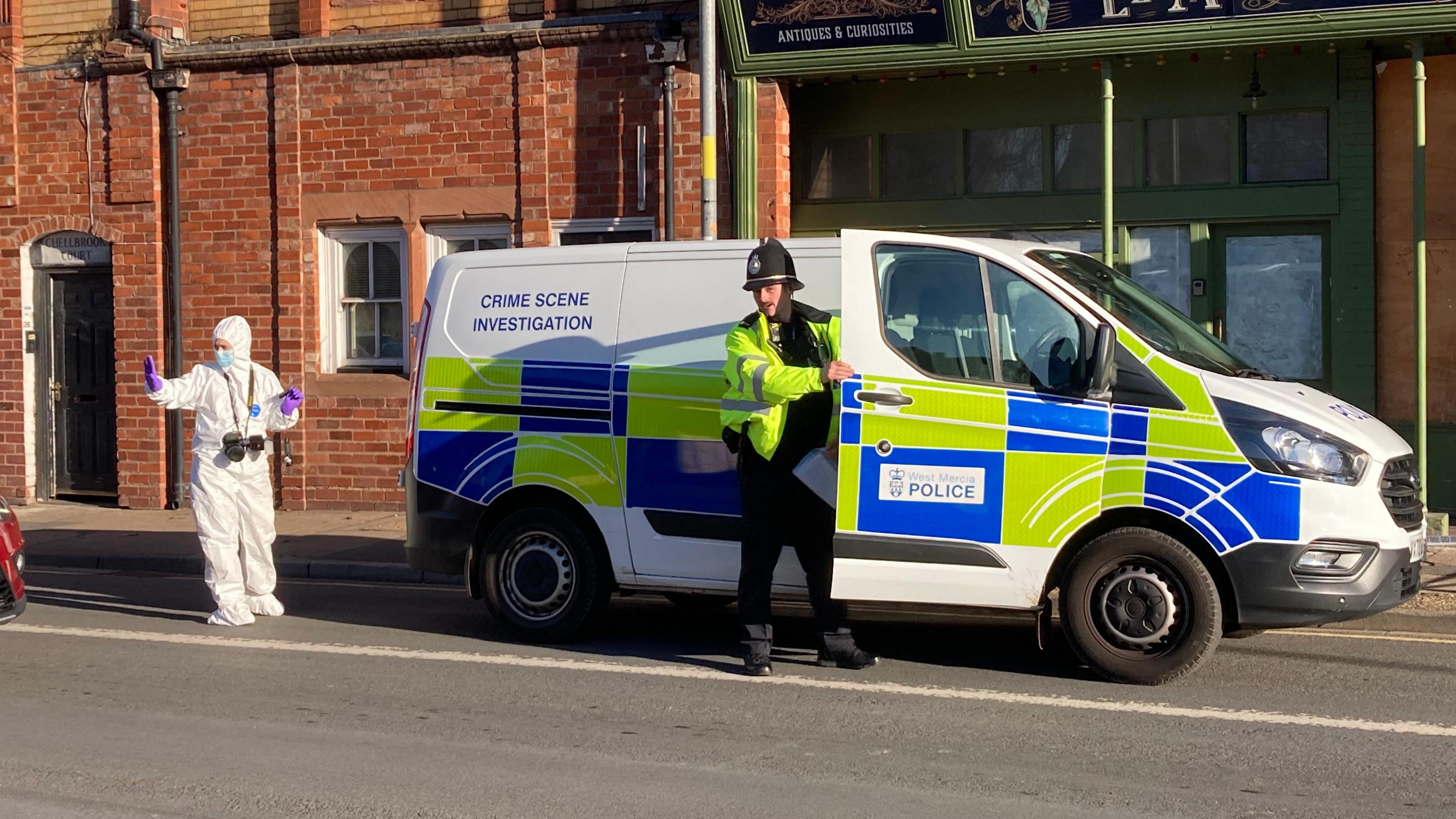 A white police van is parked outside a shop front. A police officer wearing a high viz yellow coat is seen opening the van's door while a person in a full white forensic suit holds up their hand to someone out of shot.